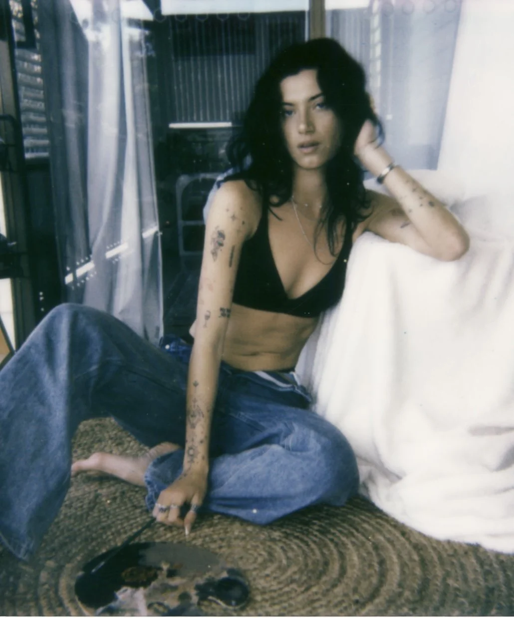 A woman with dark, wavy hair, tattoos, and a black top is sitting on a woven surface next to a white pillow, looking at the camera with a relaxed expression.