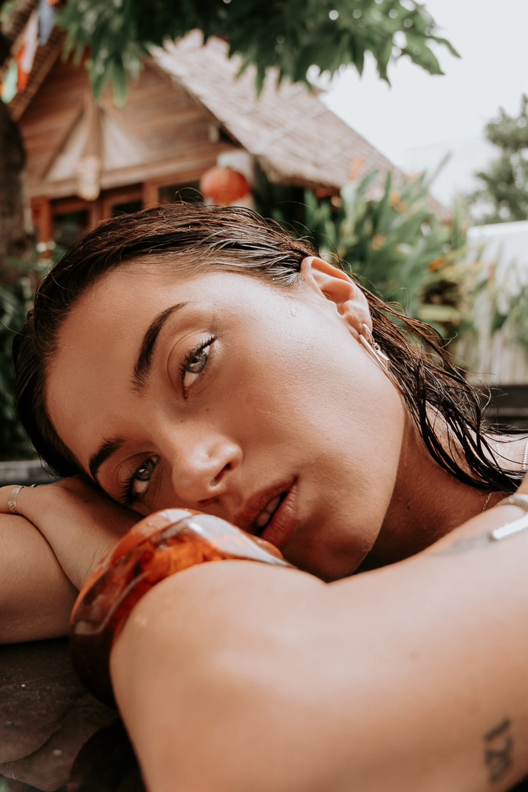 Close-up of a woman with wet hair resting her head on her arm outdoors, with lush green plants and a wooden house in the background.