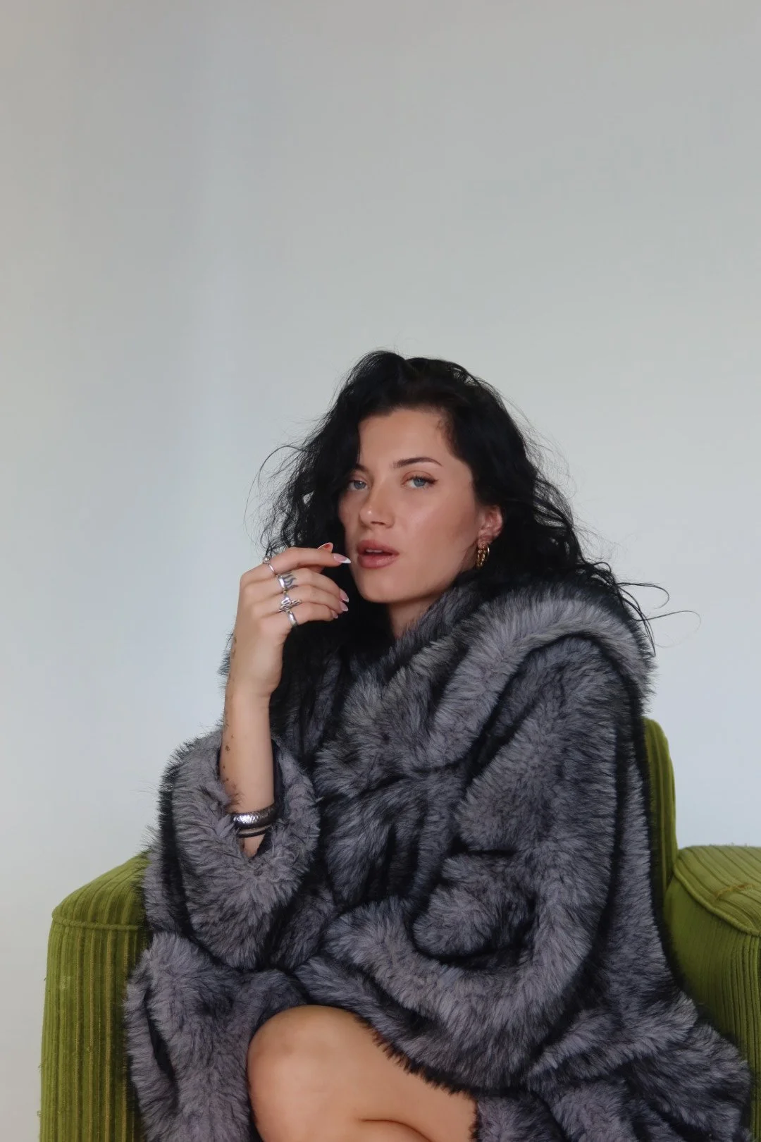 A woman with black curly hair wearing a gray faux fur coat sitting on a green chair in front of a plain white wall.