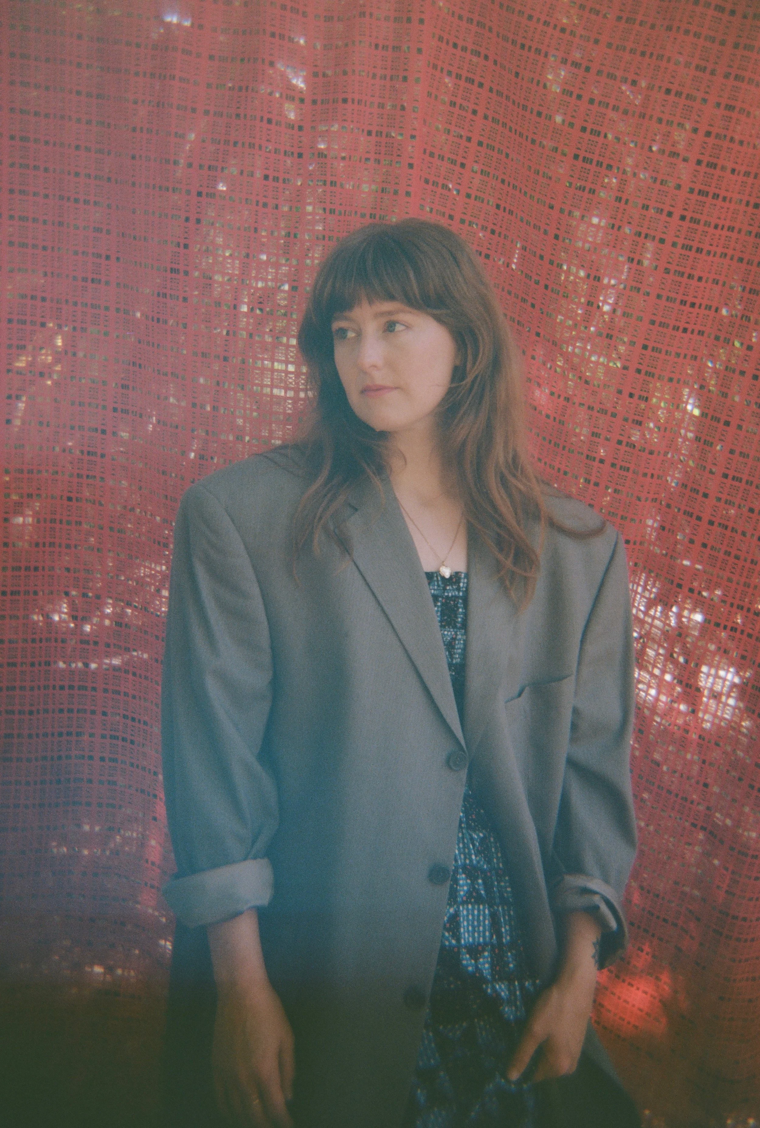 A woman with brown hair wearing a gray blazer over a patterned dress, standing in front of a red textured background.