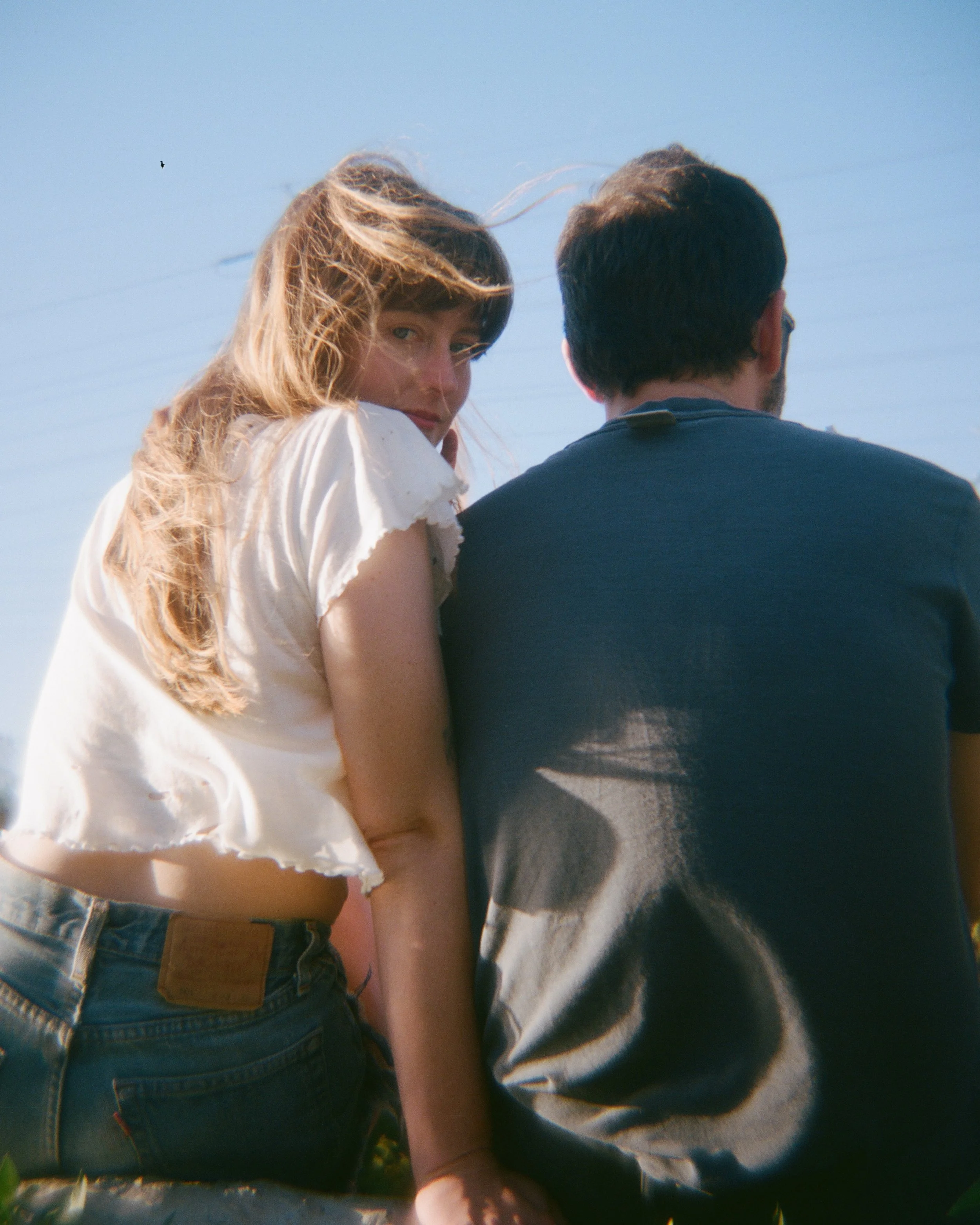 A man and a woman sitting, facing away from the camera. The woman is looking over her shoulder at the camera, against a clear blue sky.