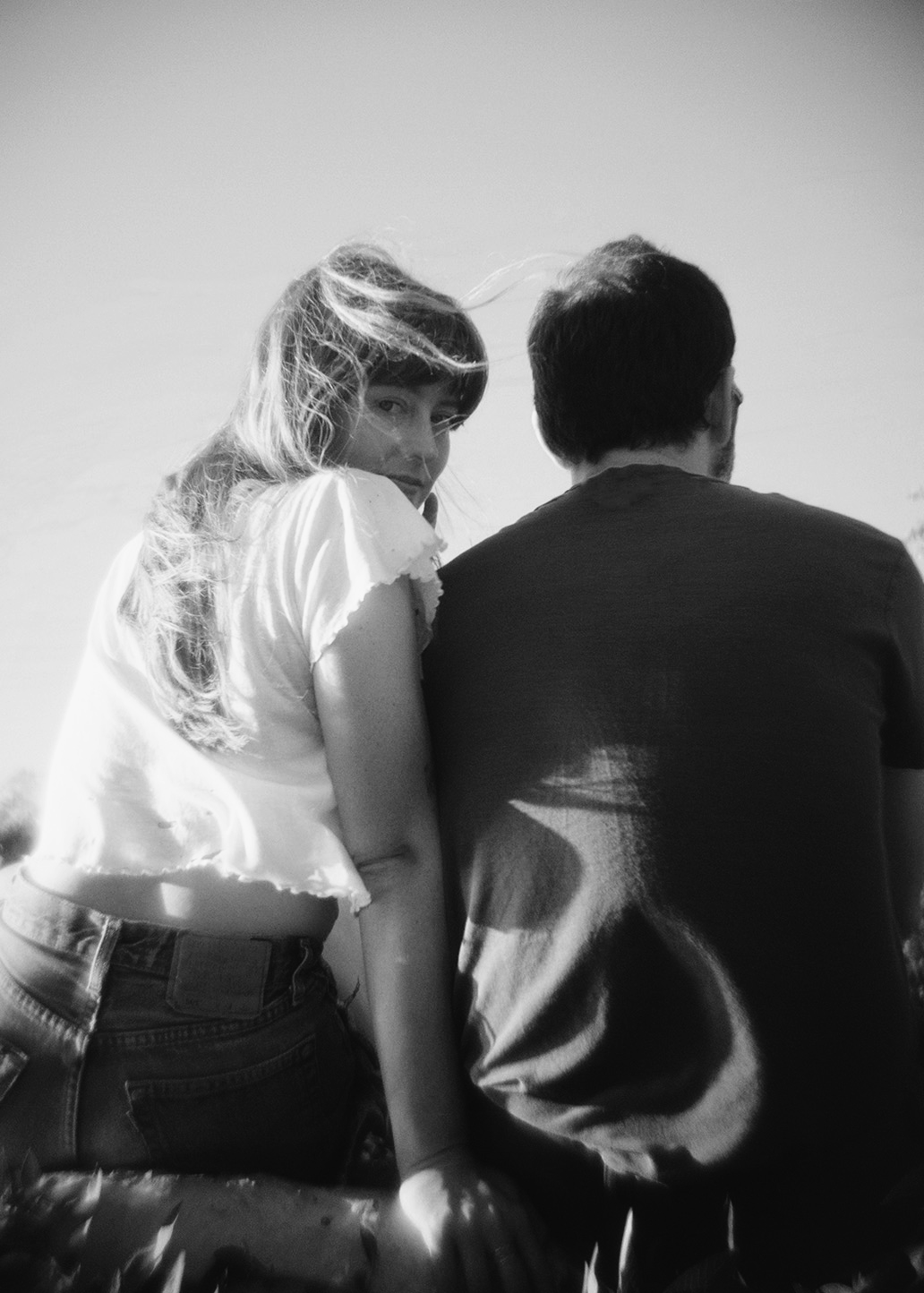 A black-and-white photo of a woman with long hair and a man sitting outdoors, with the woman looking over her shoulder.