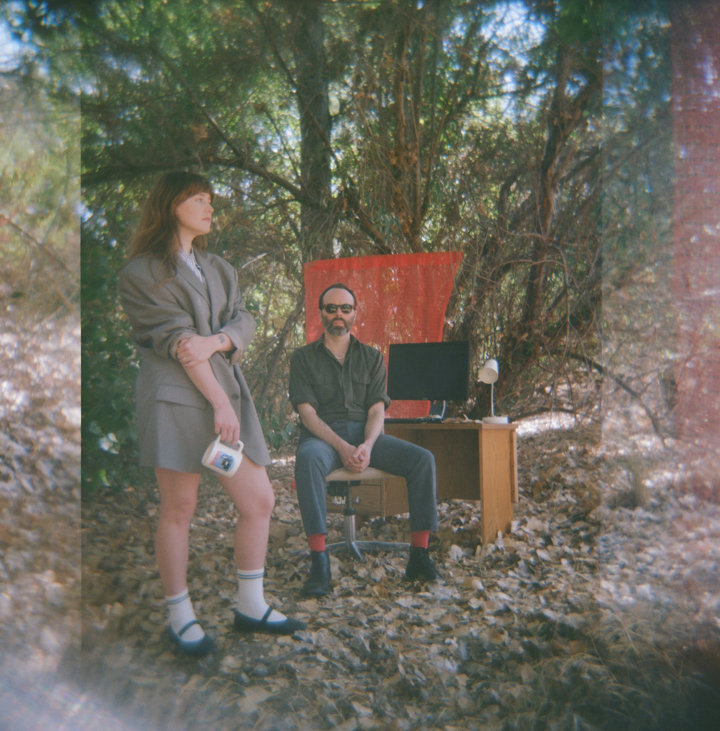Three people, a woman with long hair, a man with glasses and short hair, and a woman with glasses, sitting outdoors in a wooded area during daytime. The woman with long hair is standing, wearing a gray blazer and striped socks, holding a mug. The man
