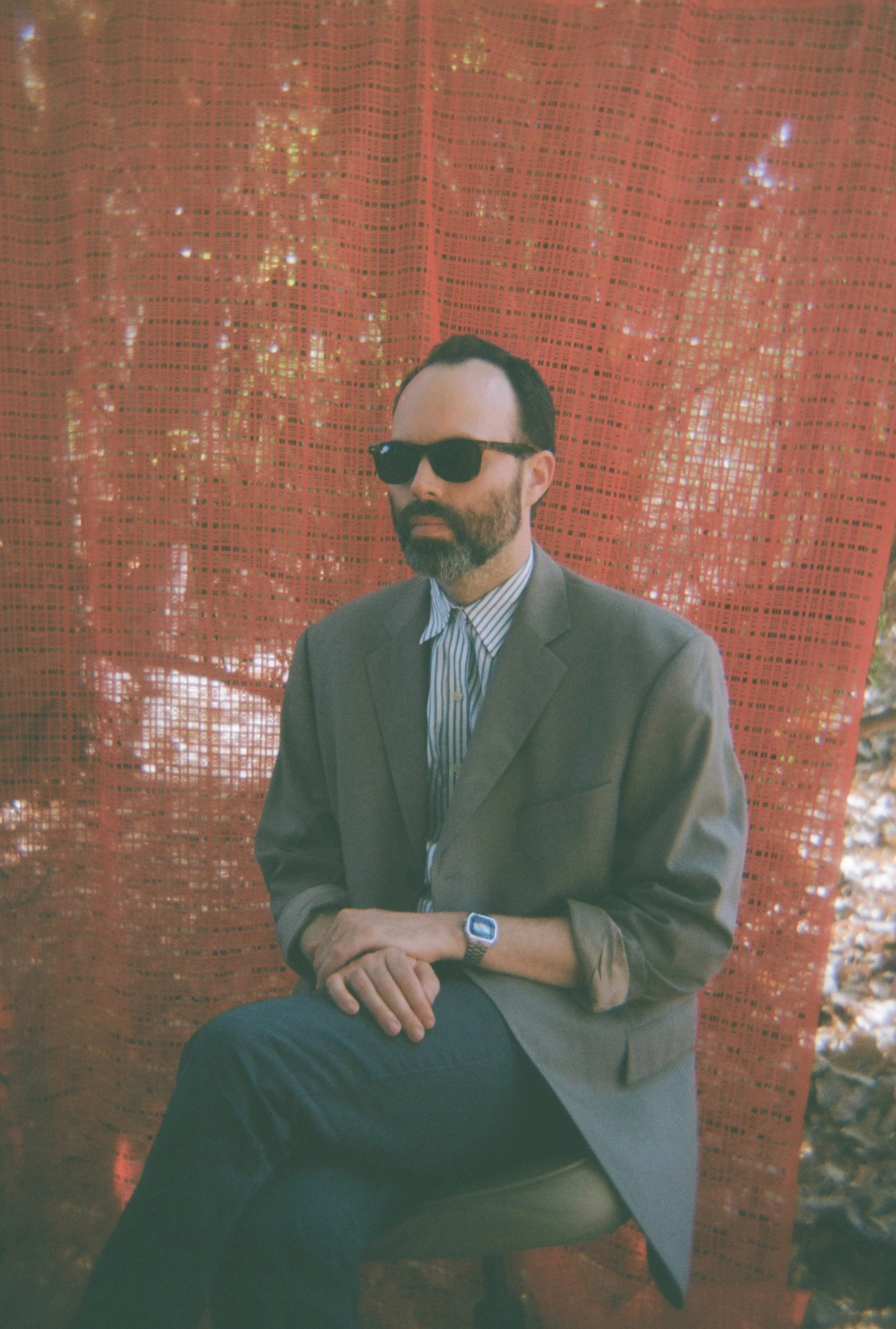 A man with dark hair, beard, dark sunglasses wearing a gray suit and striped shirt, seated with his hands clasped, in front of a red fabric background with black square patterns.