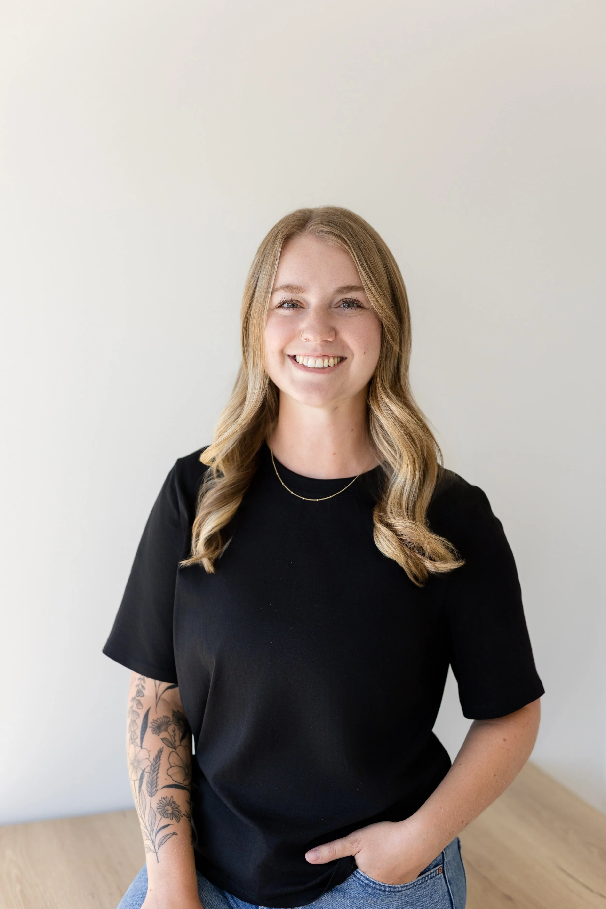 A smiling woman with blonde, wavy hair wearing a black t-shirt and blue jeans, standing against a plain light-colored background.