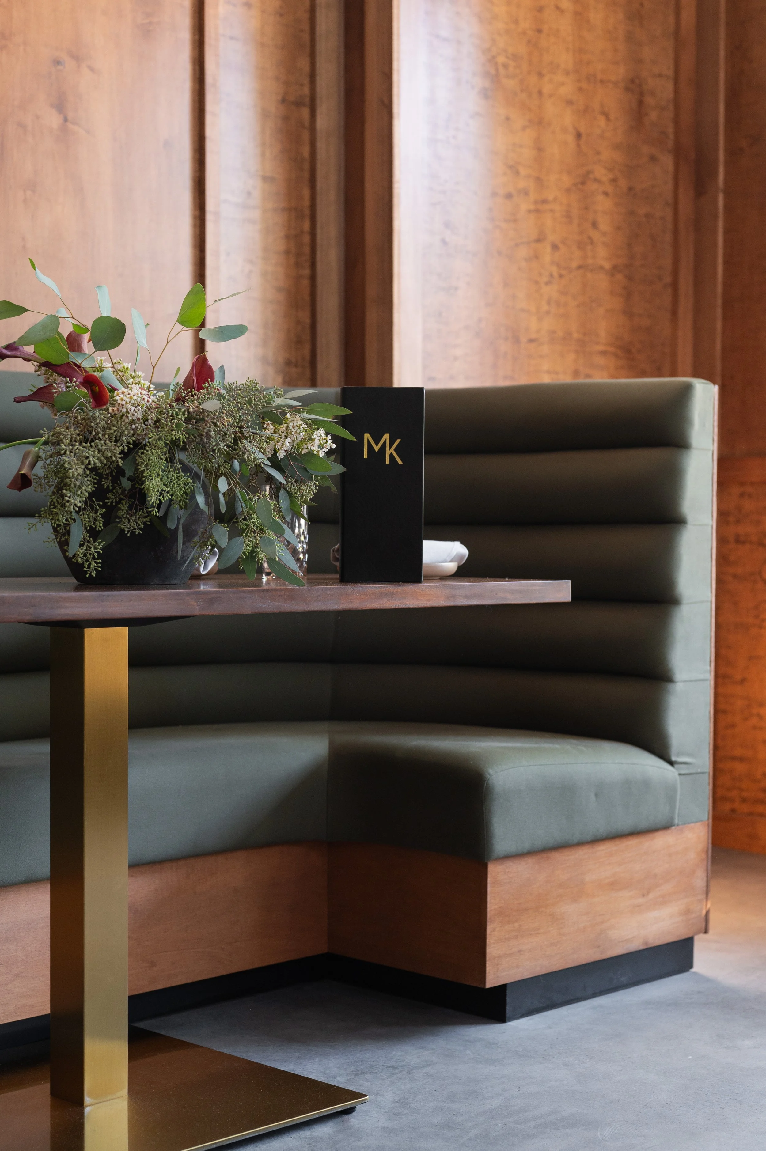 Interior of a restaurant with green upholstered banquette seating, a wooden table with a gold-colored support, a large flower arrangement in a black vase, and a black menu or card with the initials 'MK' on it, against wooden wall paneling.