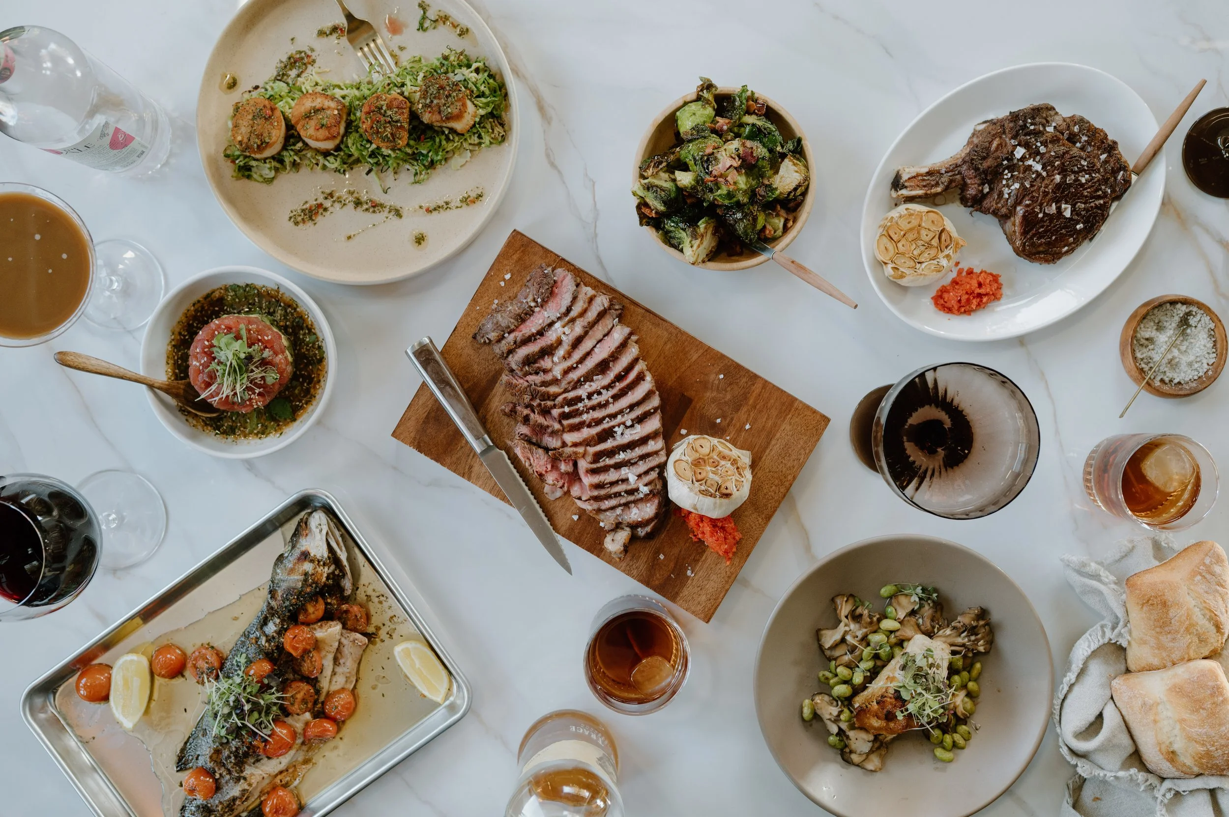 A table with various dishes including sliced steak on a wooden cutting board, a salad with Brussels sprouts, a bowl of tomato soup, a plate with a cooked fish garnished with cherry tomatoes, and various other small bowls of appetizers and drinks.
