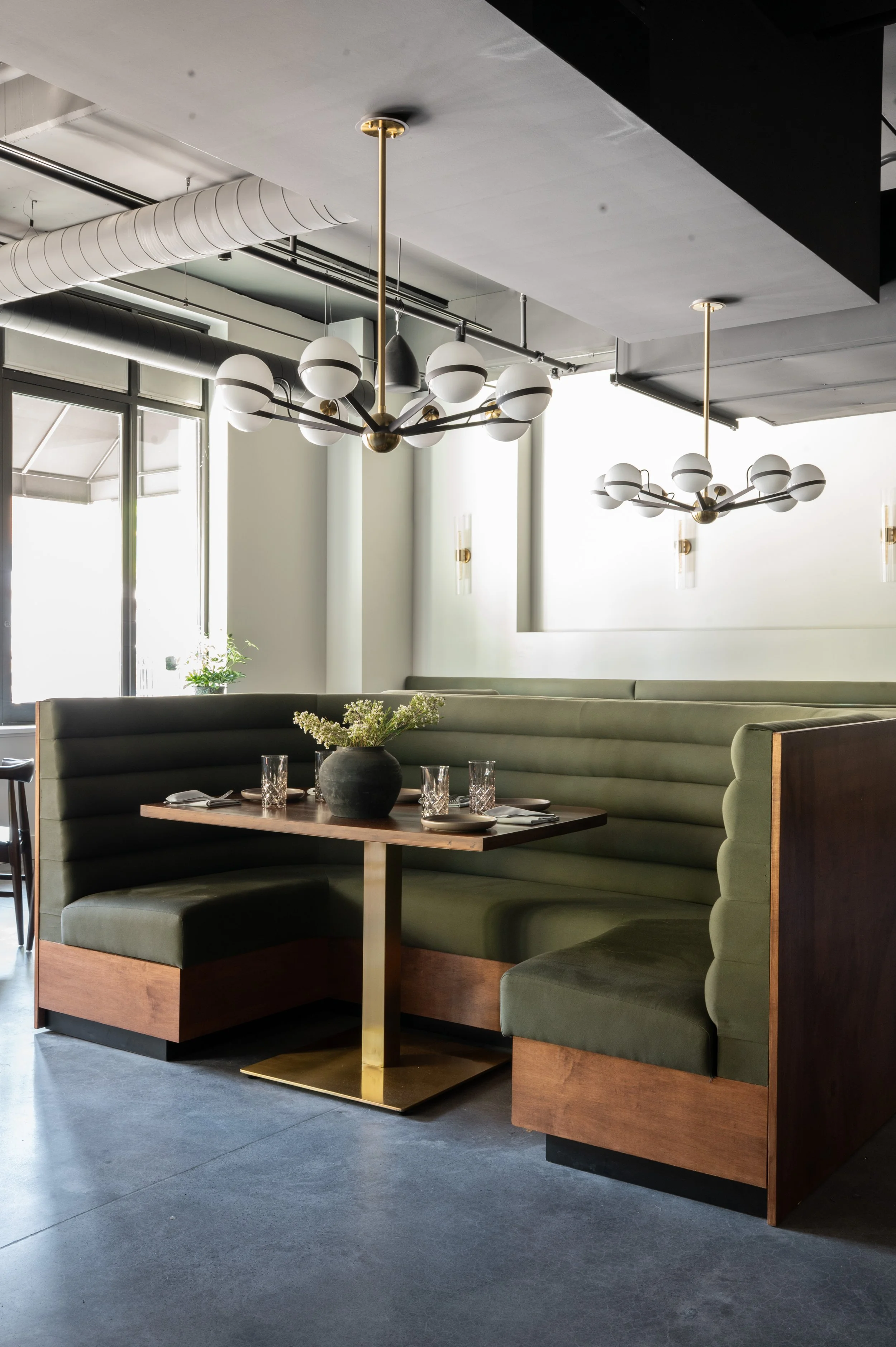 Modern restaurant booth with green upholstery, wooden base, and a table set with glasses, plates, and a black flower vase, illuminated by contemporary ceiling and wall lights.