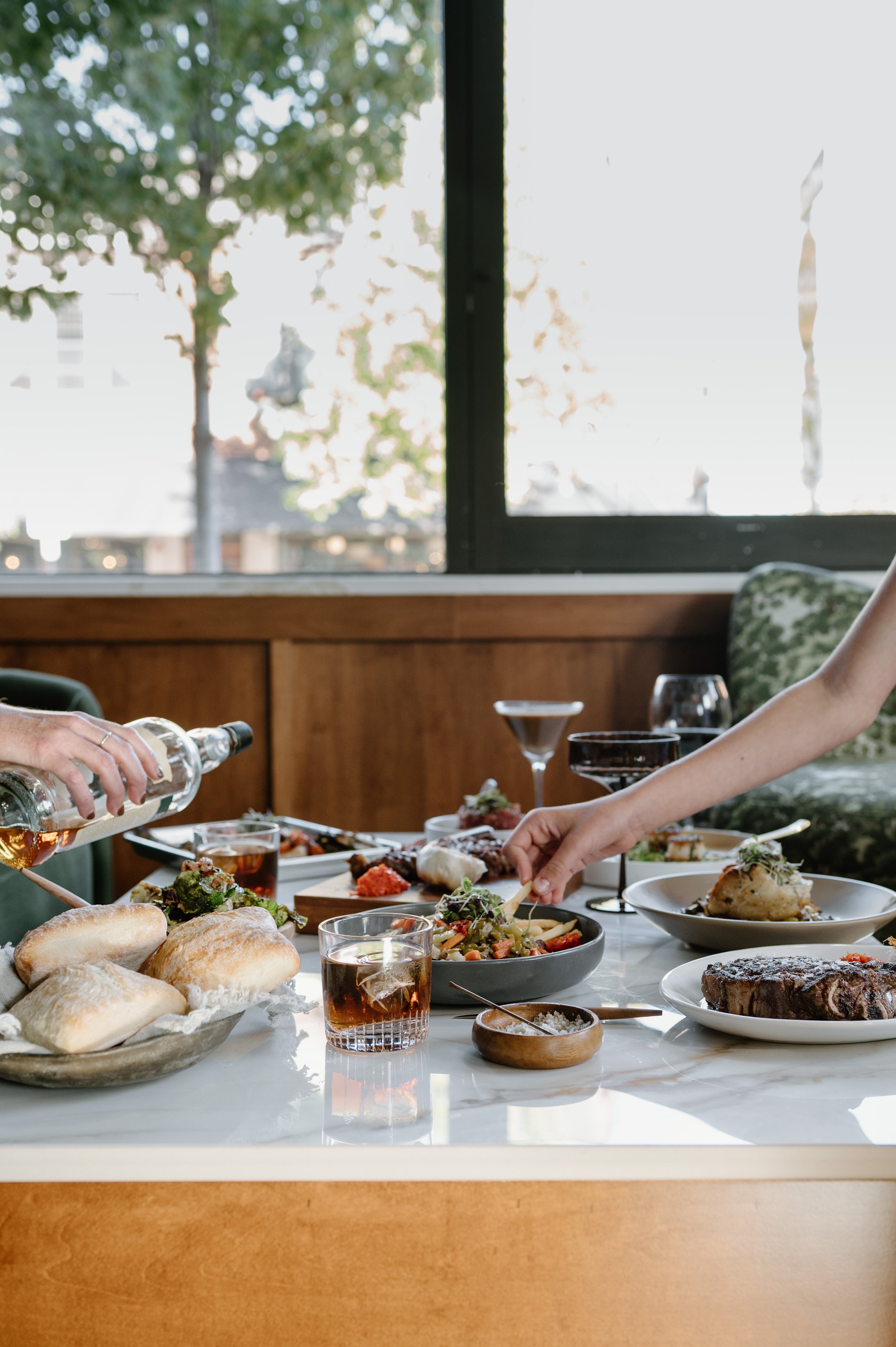 A table set for a meal with various dishes, including bread rolls, salads, a steak, and drinks like wine and cocktails. There are several hands reaching for food and pouring drinks.