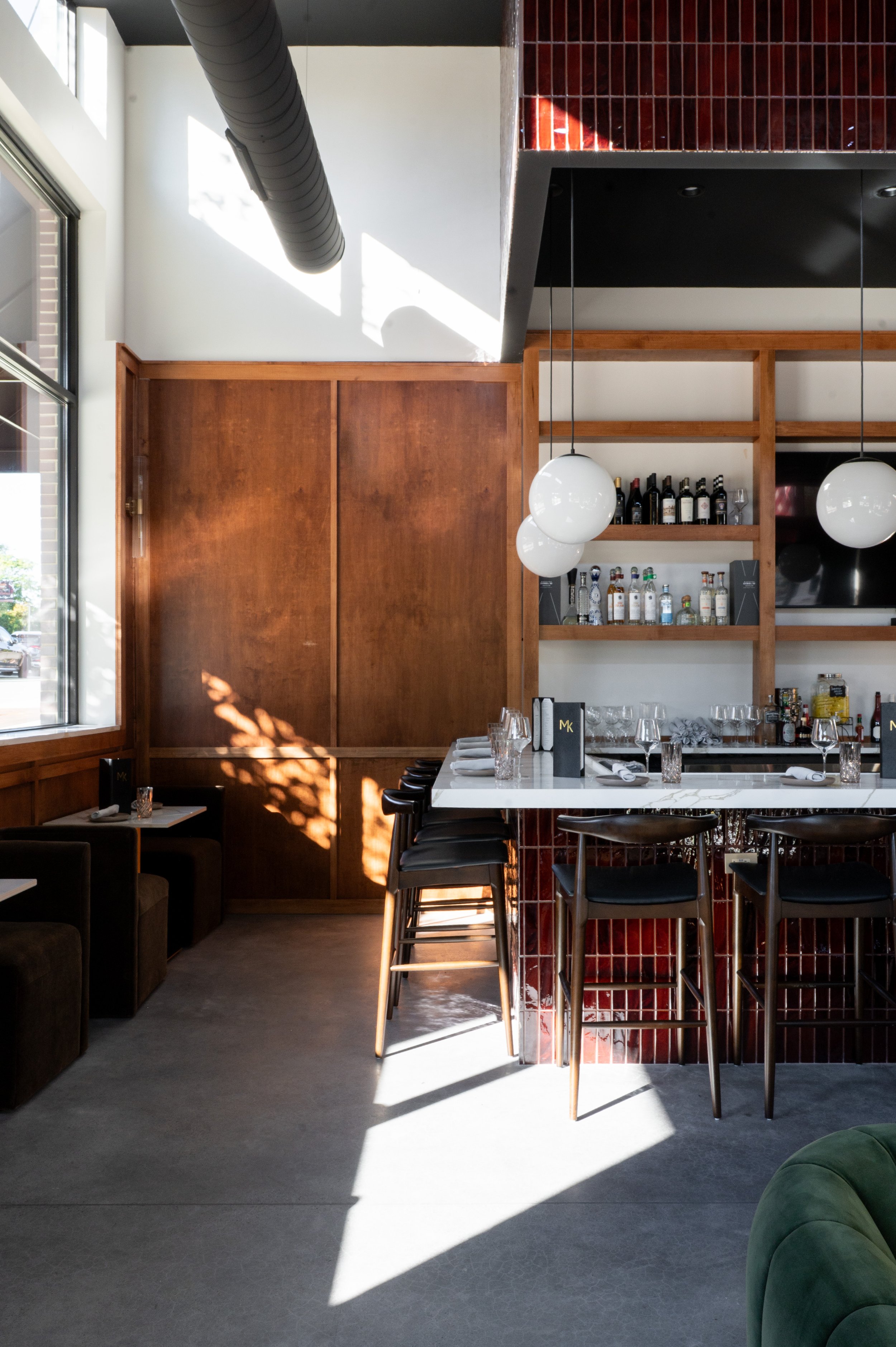 Interior of a modern restaurant or bar with a white marble counter, black chairs, hanging white globe lights, wooden shelves with bottles, and a large window letting in natural light.