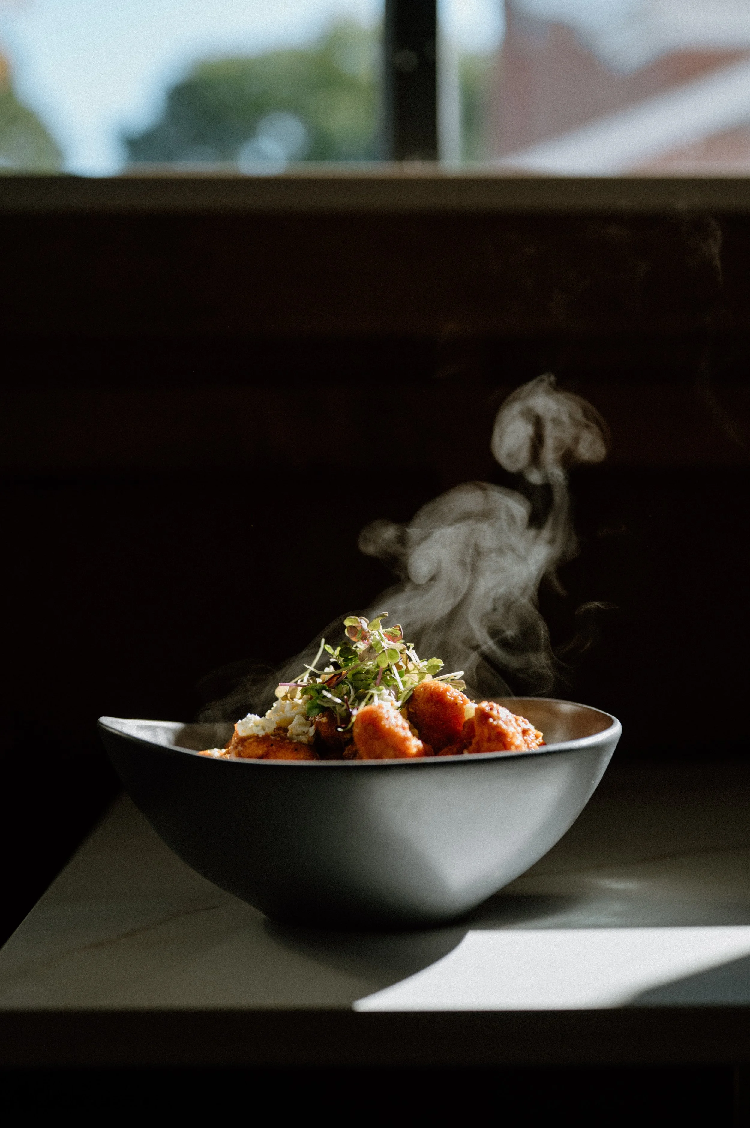 A steaming bowl of food, possibly fried chicken or similar, garnished with microgreens and cheese, on a white surface near a window with natural light.
