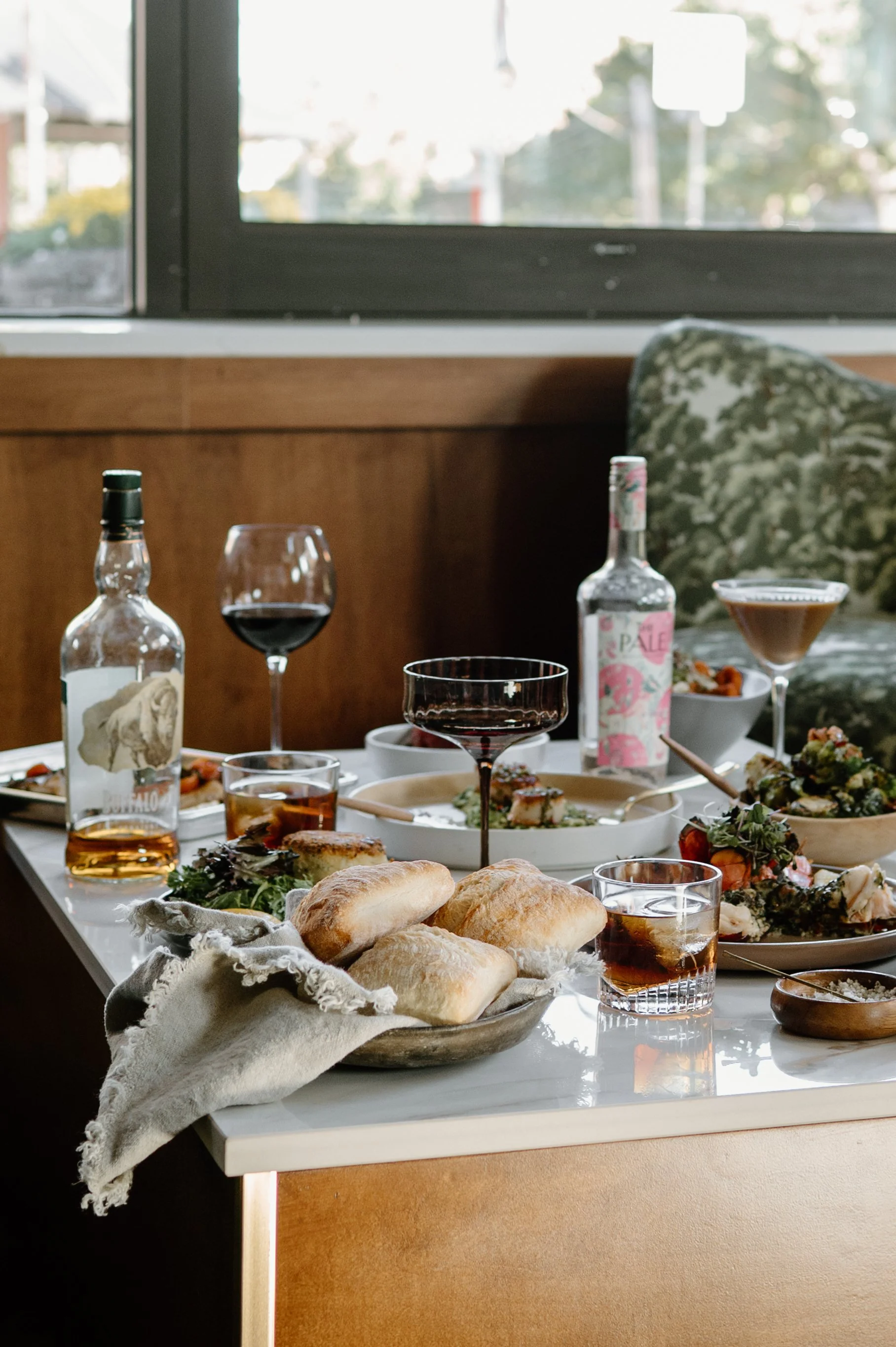 A table set with various dishes, glasses of red wine, bottles, and bread in a cozy dining area with a window in the background.