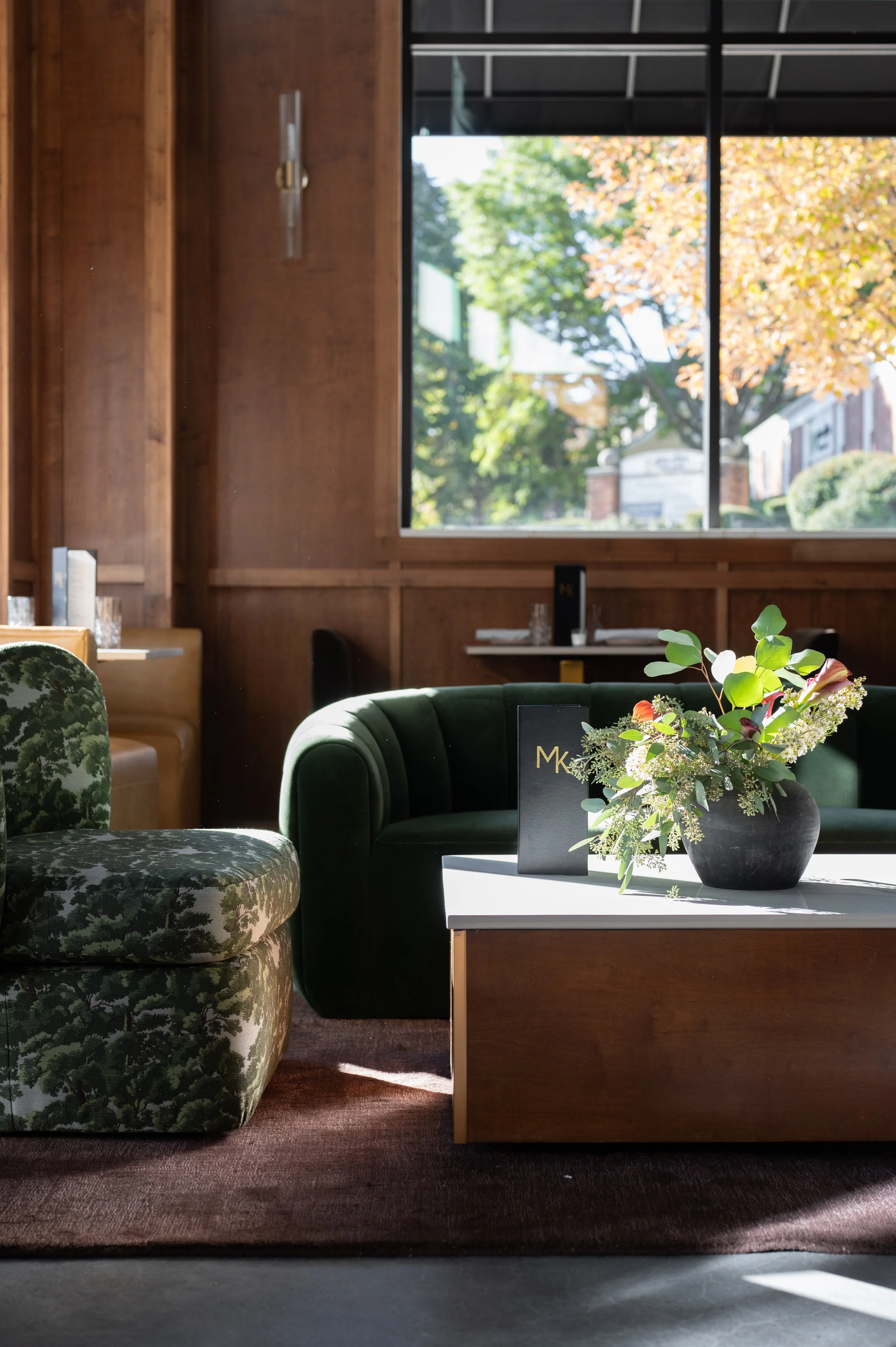 Interior of a cozy, stylish lounge with a green velvet sofa, floral patterned armchair, wooden walls, a large window showing trees outside, and a white table with a black vase holding greenery and flowers, along with a menu.