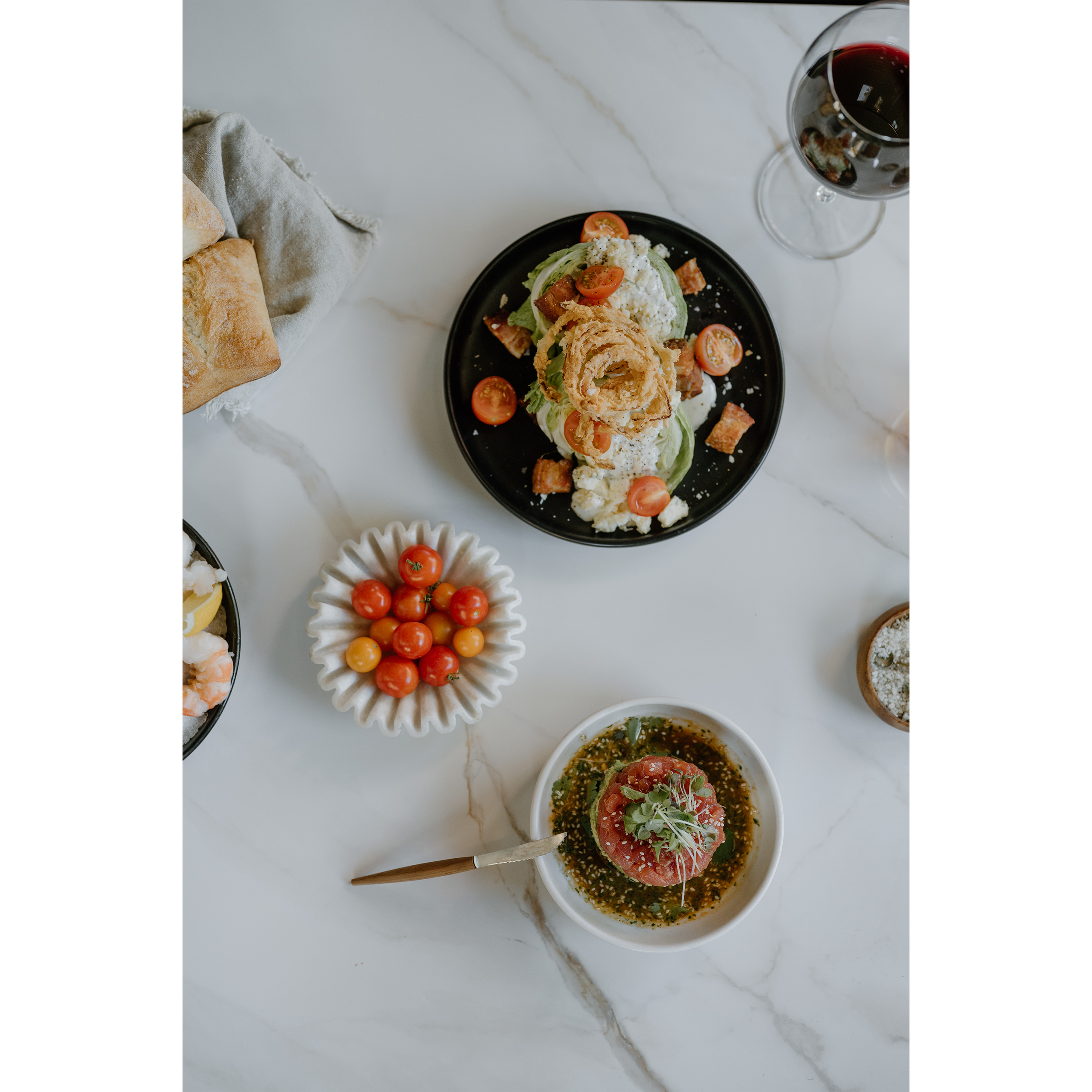 A dinner table with a black plate of salad topped with fried onion rings, cherry tomatoes, croutons, and cheese, a bowl of cherry tomatoes, a bowl of seaweed salad garnished with microgreens, a glass of red wine, and bread on a marble surface.