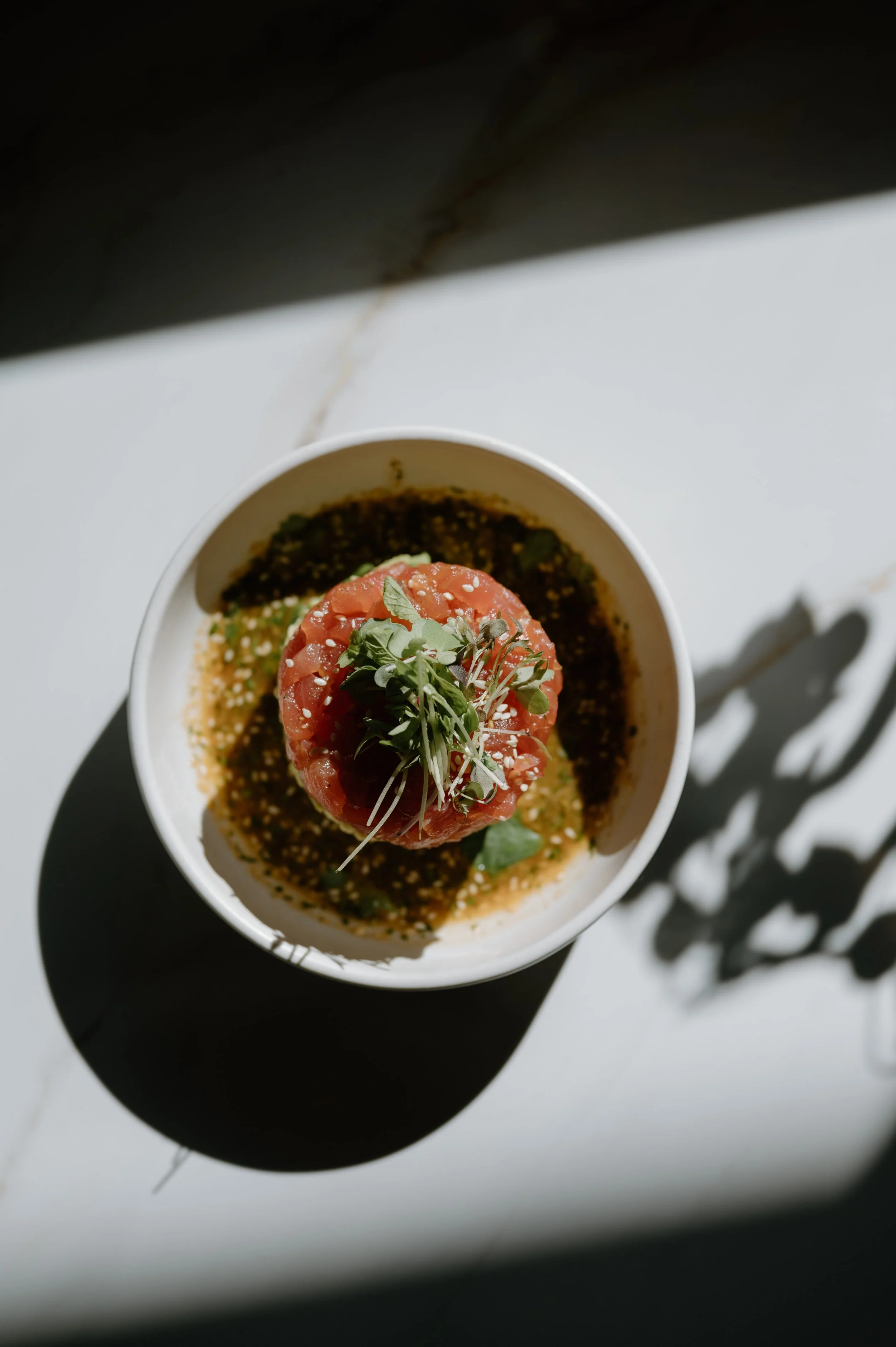 A bowl with a halved tomato topped with microgreens, garnished with sesame seeds, surrounded by a dark green sauce or dressing, on a white surface with shadows and light.
