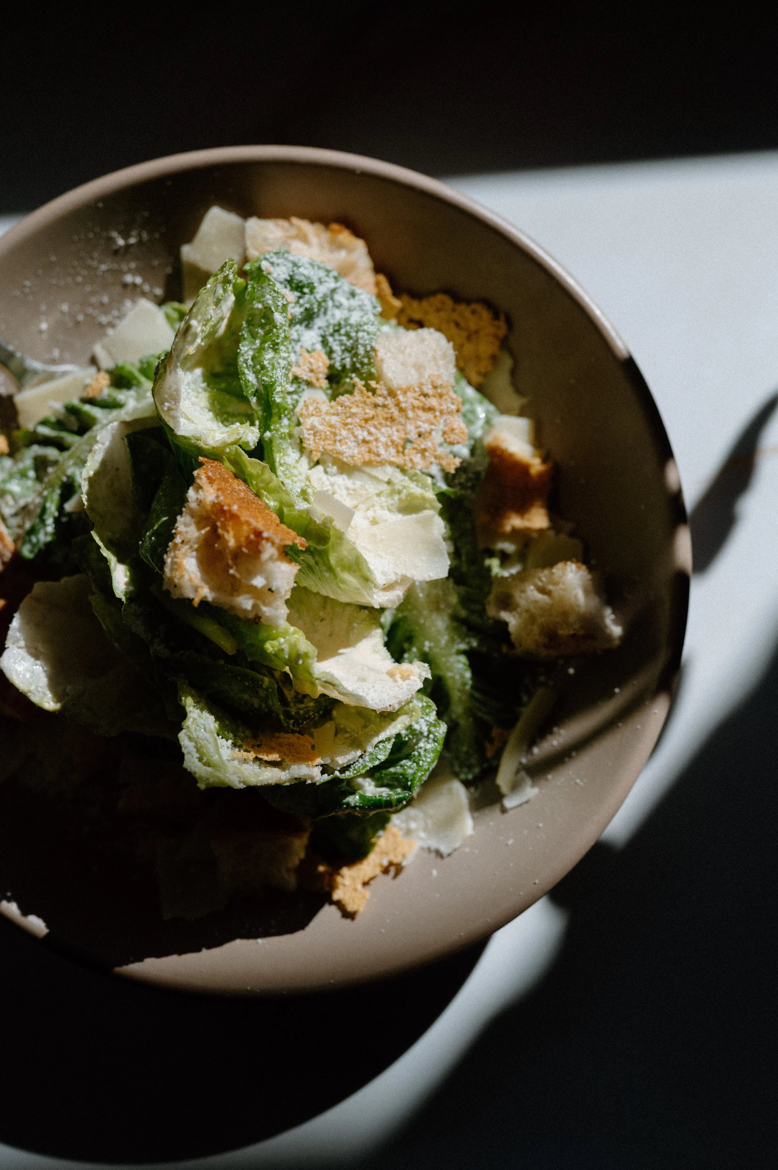 Close-up of a Caesar salad in a beige bowl, with romaine lettuce, croutons, Parmesan cheese, and Caesar dressing, partially illuminated by sunlight.