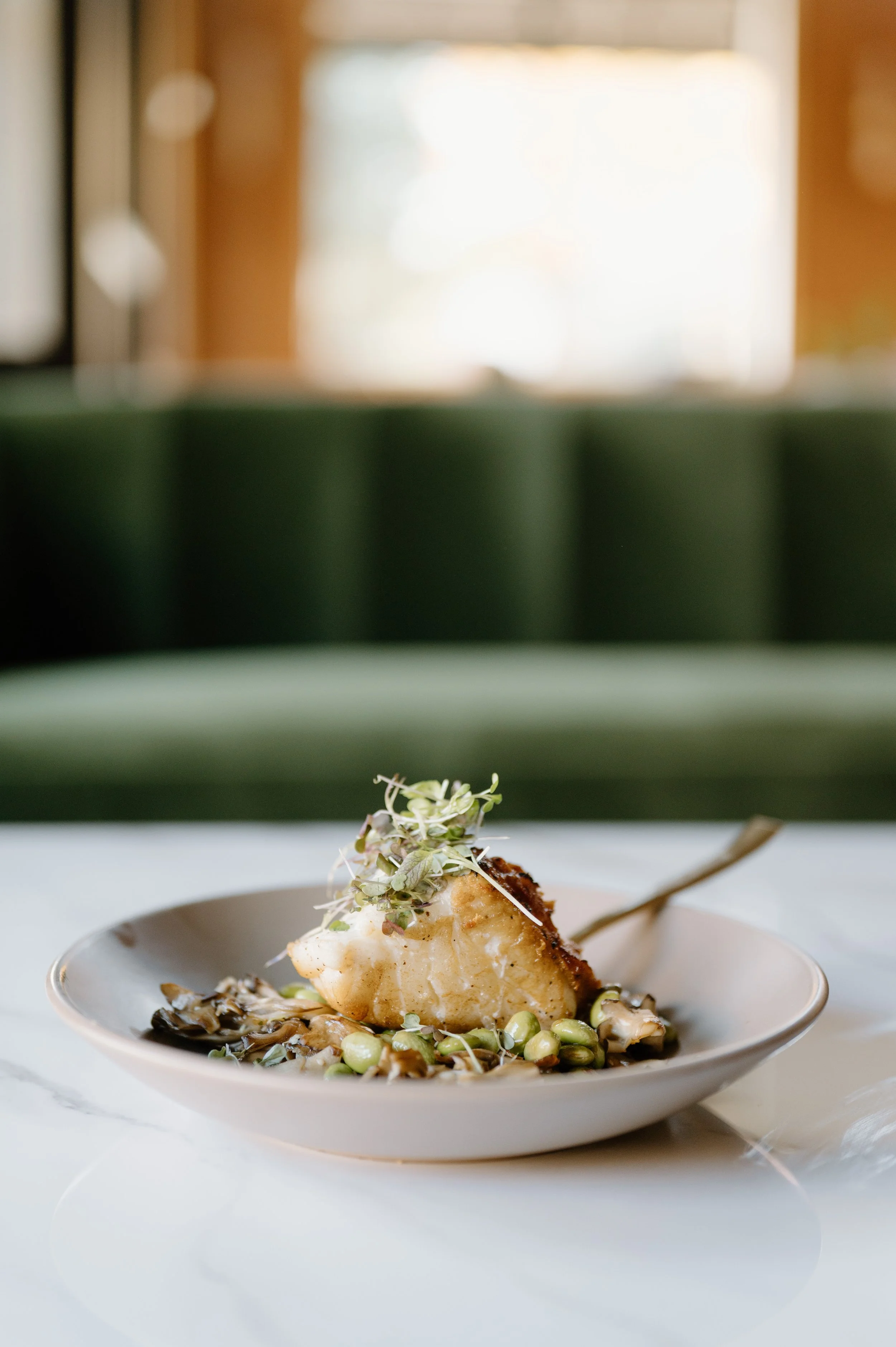 A plated dish featuring a cooked fish fillet topped with microgreens, served with roasted vegetables and beans on a white plate.