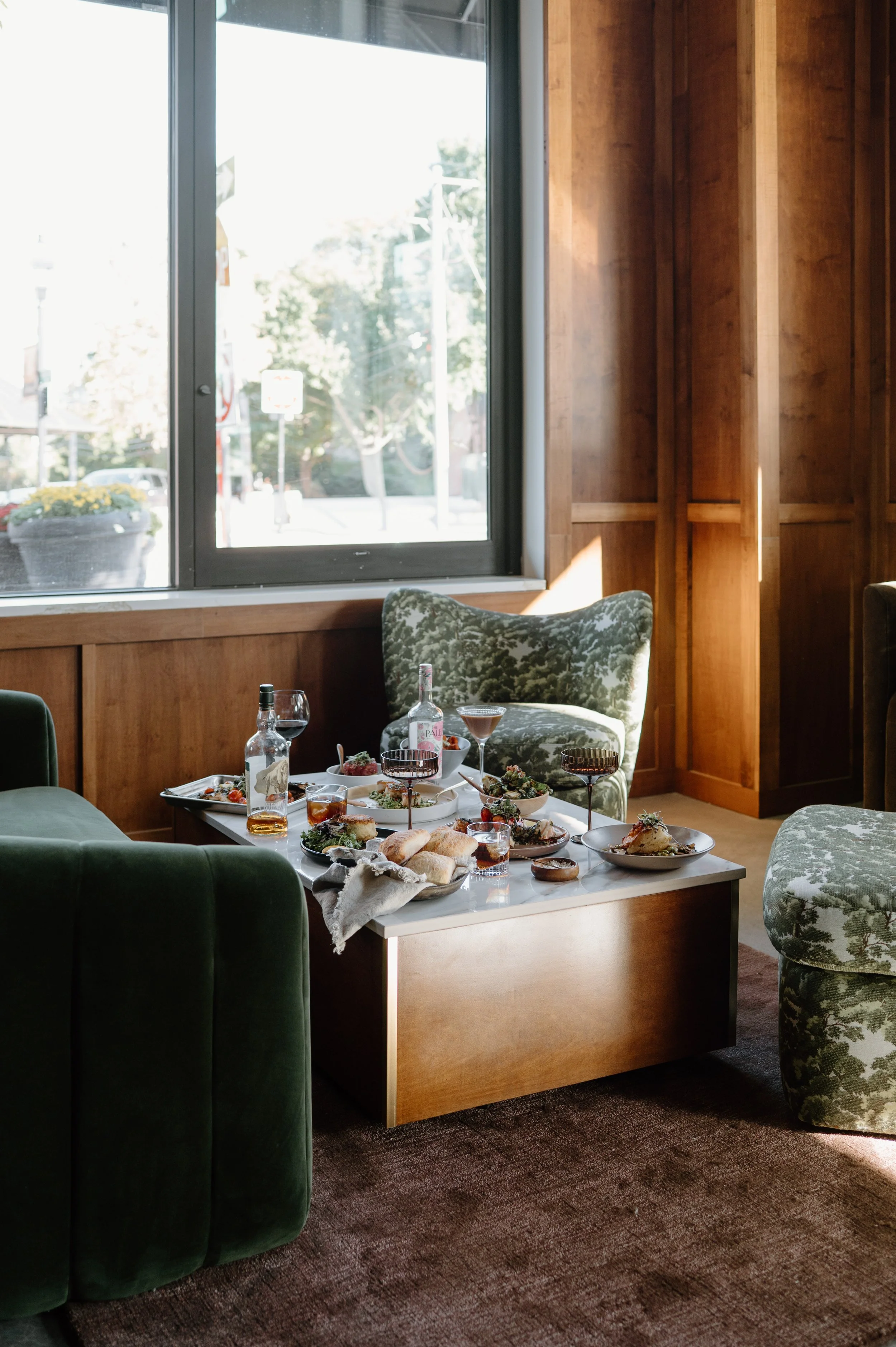 A table set with food and drinks in a cozy room with large window and green velvet furniture.