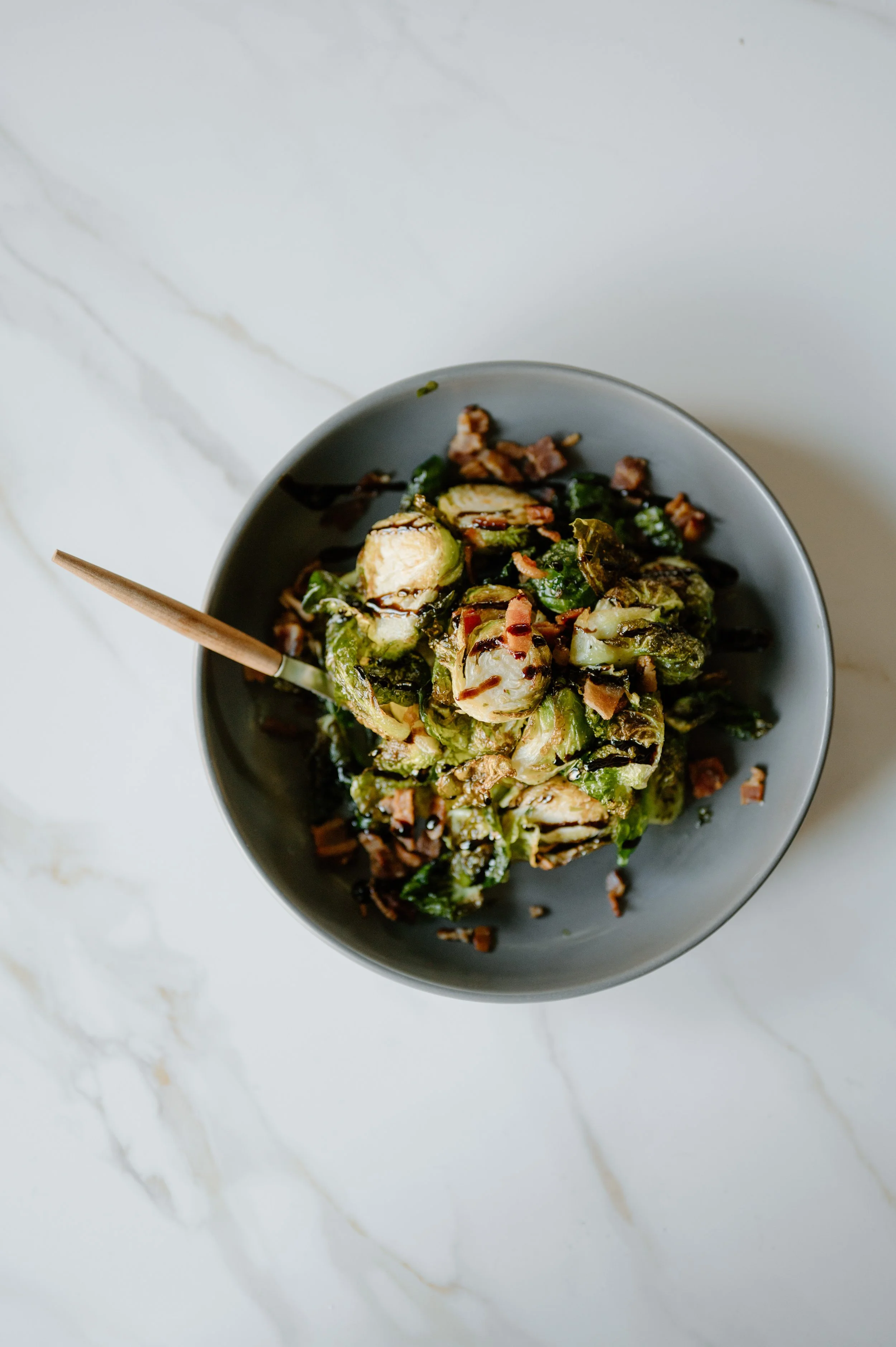A gray bowl containing roasted Brussels sprouts with bacon bits on a white marble countertop.