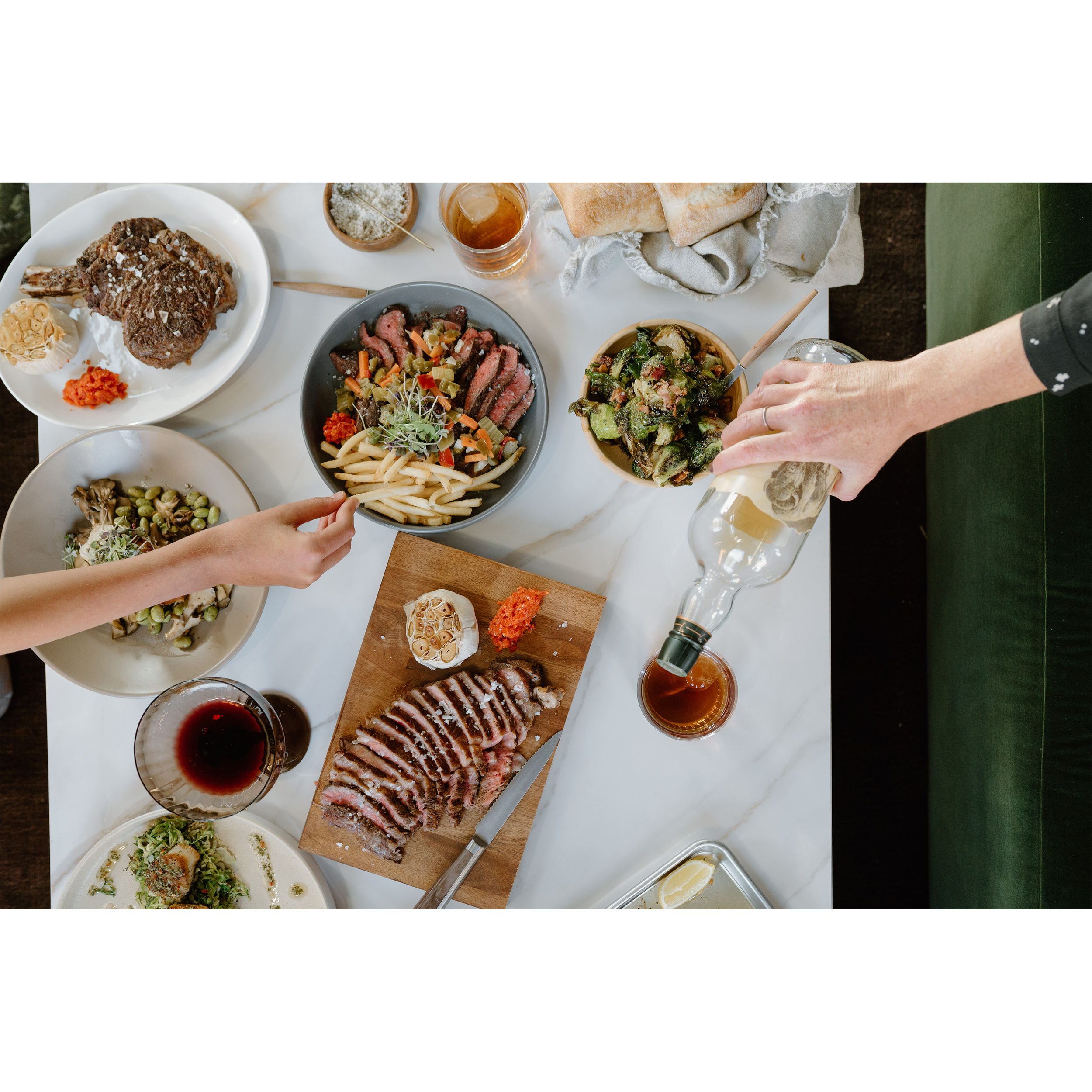 Overhead view of a table with various dishes including sliced steak, Brussels sprouts, pasta, bread, roast meat, wine, beer, and salads, with people serving and sharing food.