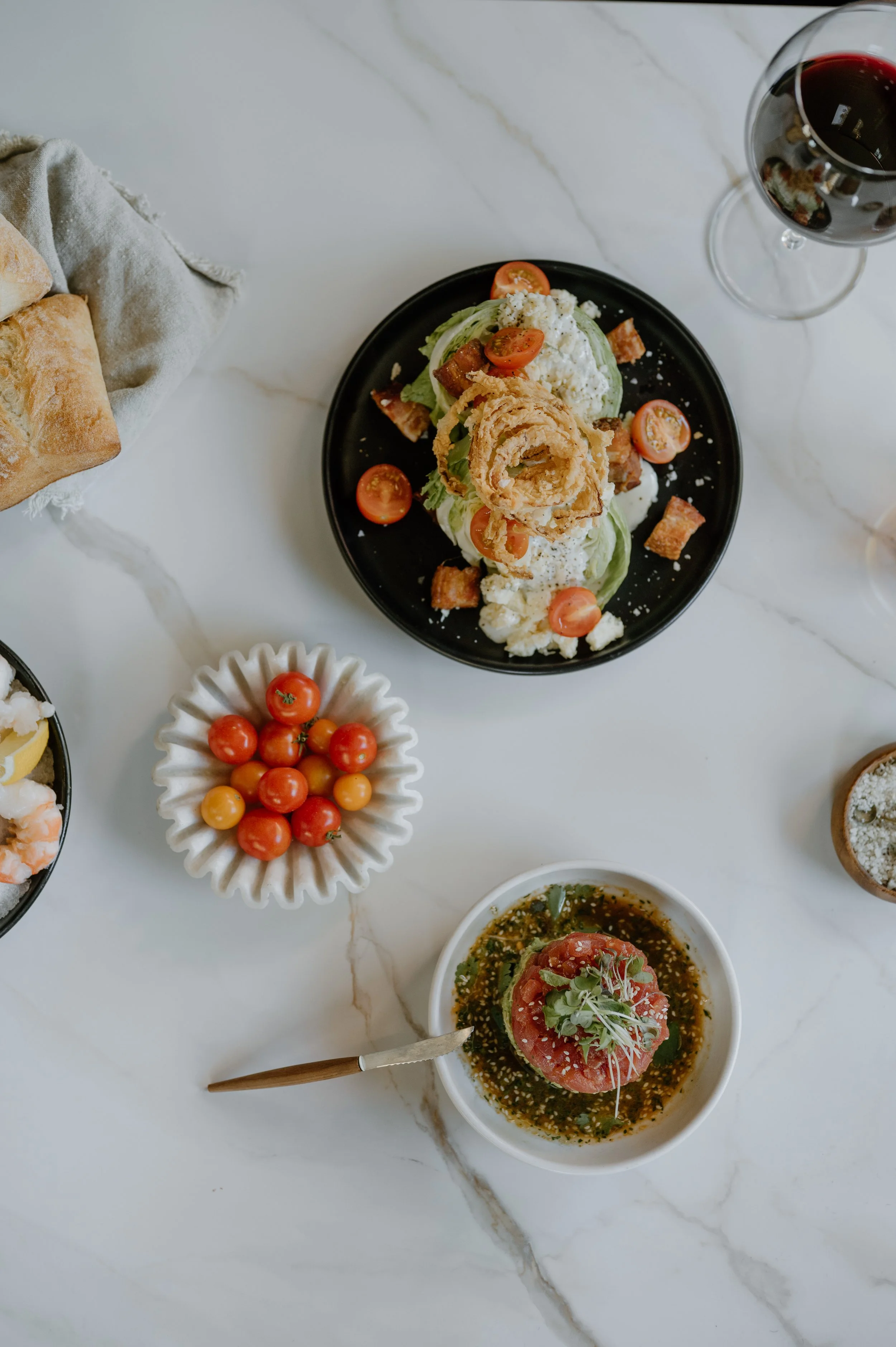 Salad with cherry tomatoes, fried onions, bacon bits, and lettuce on a black plate, a bowl of cherry tomatoes, a glass of red wine, and a dish with a tomato and basil topping on a white marble table.