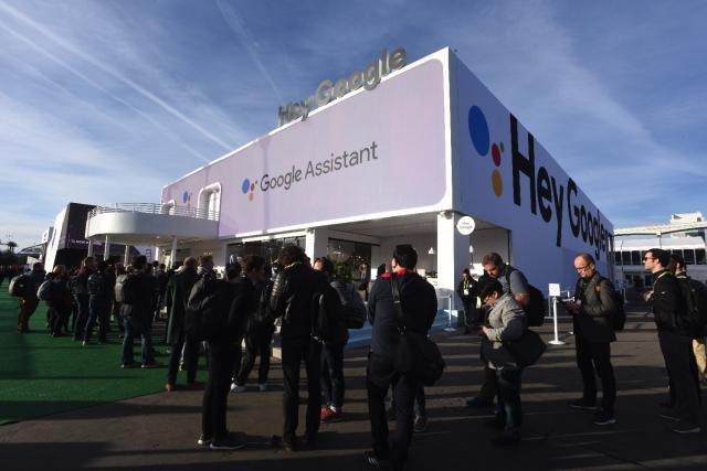 People gathered outside a large white building with 'Hey Google' and 'Google Assistant' signage under a blue sky.