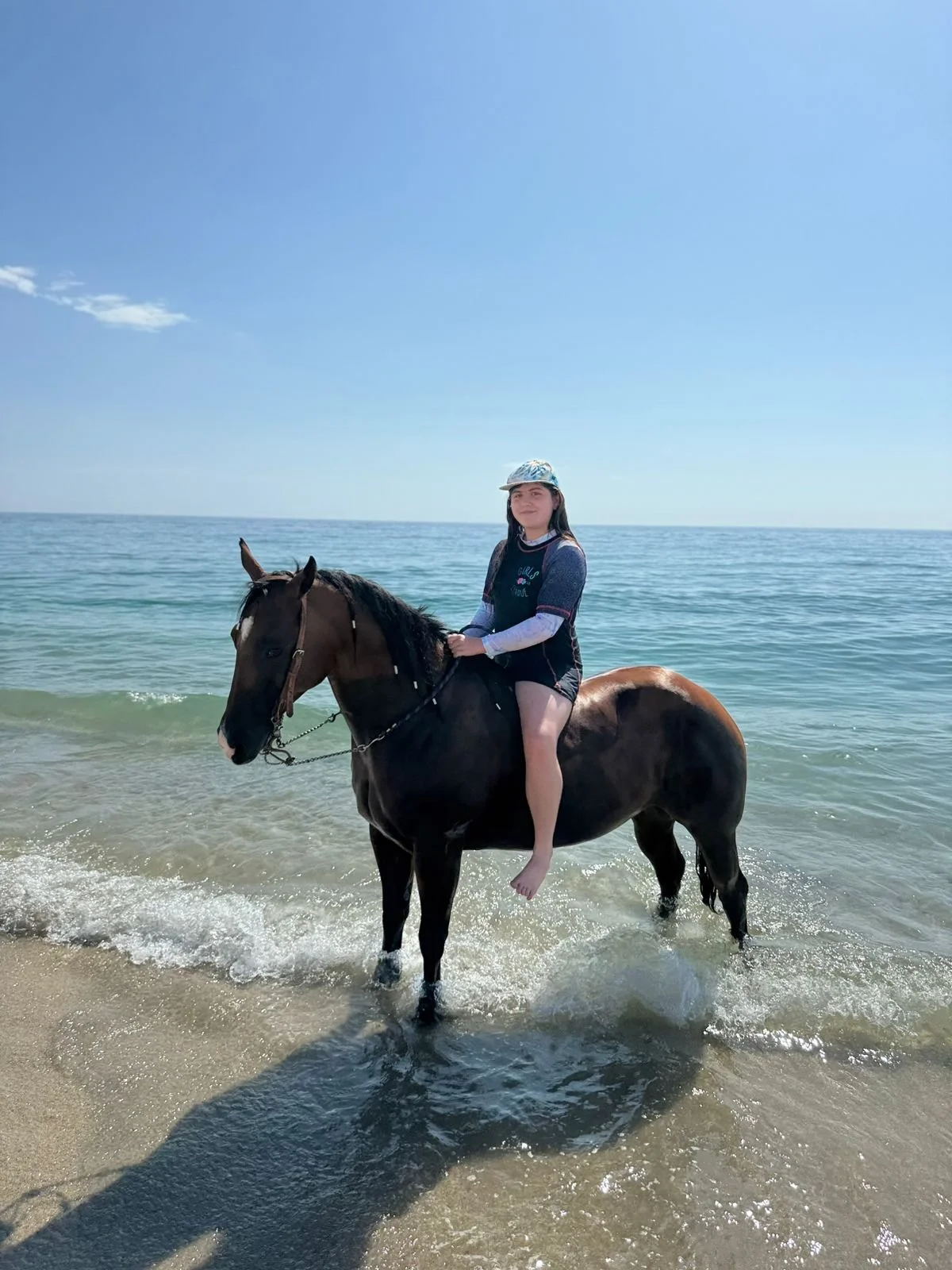 A girl riding a horse in the ocean water with a sandy beach and clear blue sky in the background.