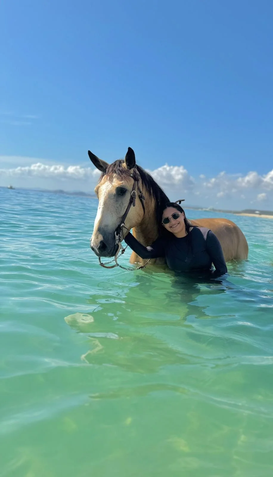 A woman in black clothing and sunglasses hugging a brown and white horse in shallow green water, with a blue sky and a distant shoreline in the background.