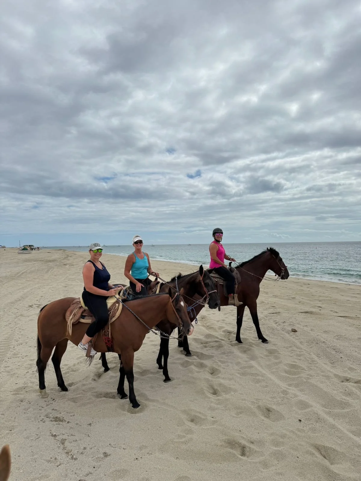 Three women riding horses on the beach under a cloudy sky.