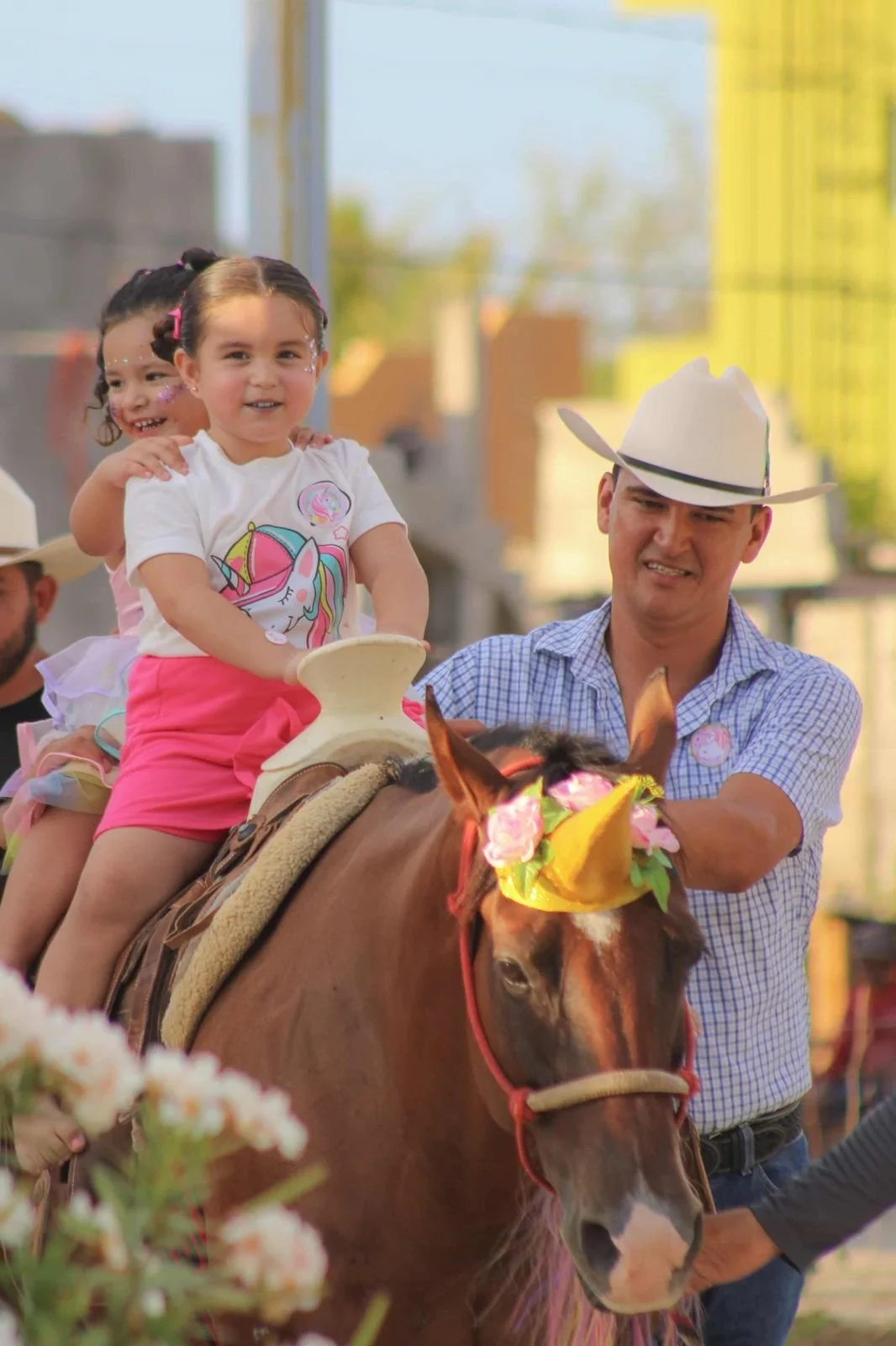 Children riding a horse at a festive event, with a man guiding the horse, and decorated with colorful accessories and a hat.