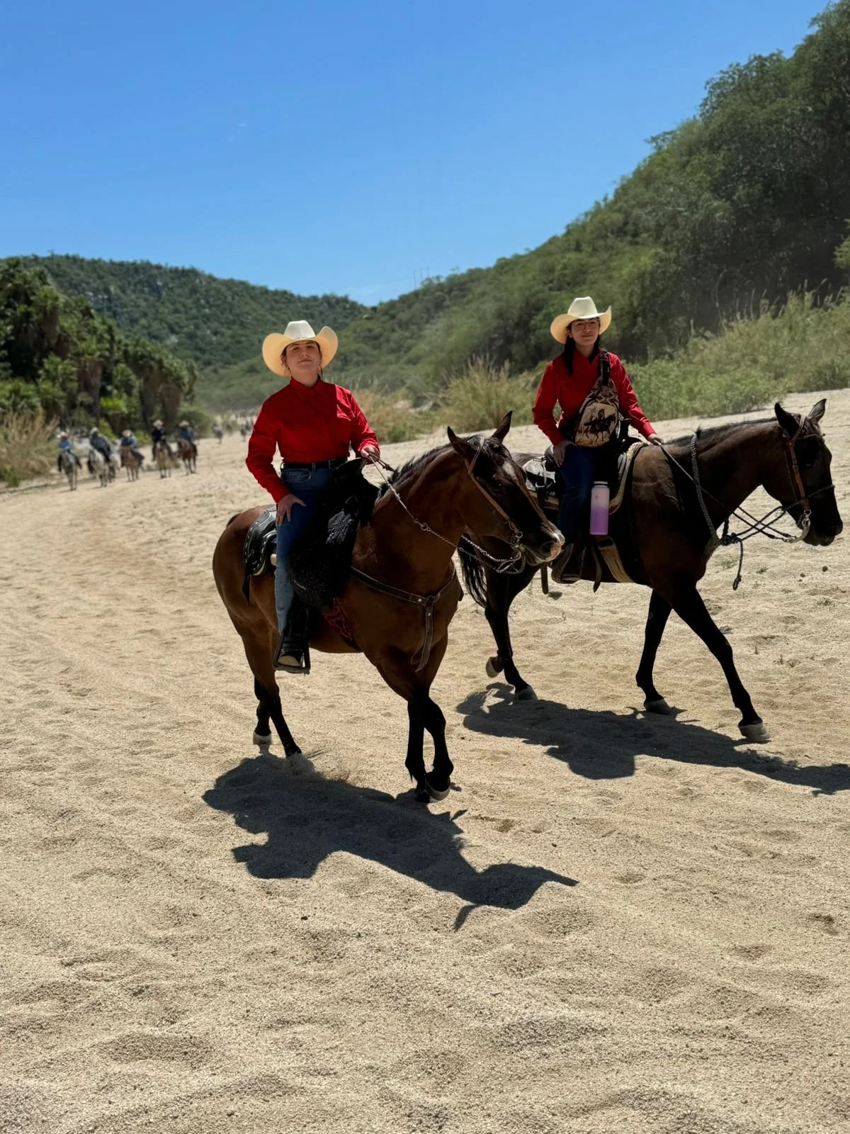 Two women riding horses on a sandy trail through a green hillside, both wearing cowboy hats and red shirts, with a group of riders visible in the background and clear blue sky overhead.