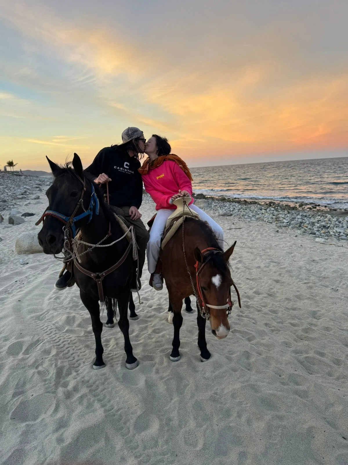 Two women kissing while riding horses on a beach at sunset, with a calm ocean and rocks in the background.