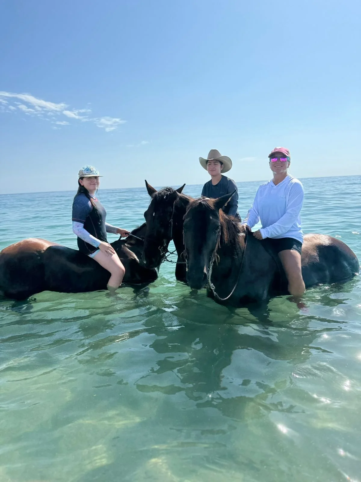 Three people riding horses in the water at the beach with clear blue sky above.
