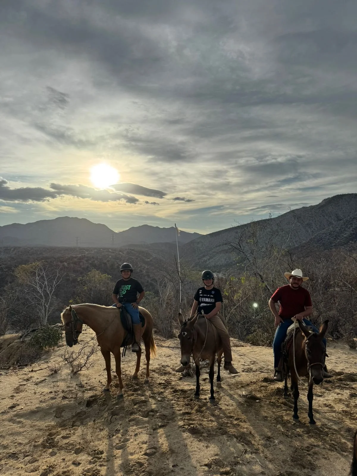 Three people riding horses on a trail in a mountainous area during sunset or sunrise with cloudy sky.