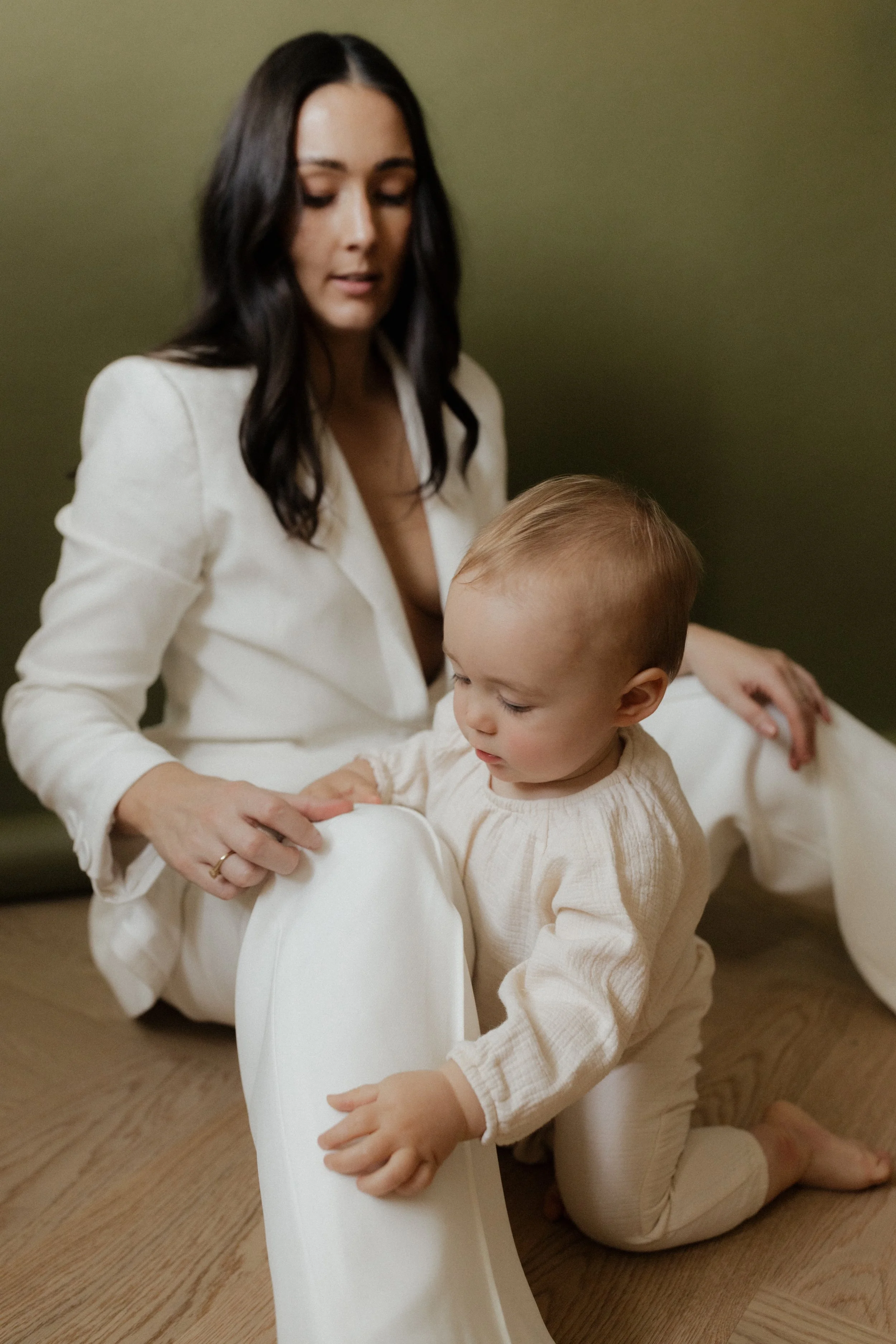 A woman with dark hair and a young child with blond hair sitting on a wooden floor, both dressed in light-colored clothing, with the woman holding the child's foot.