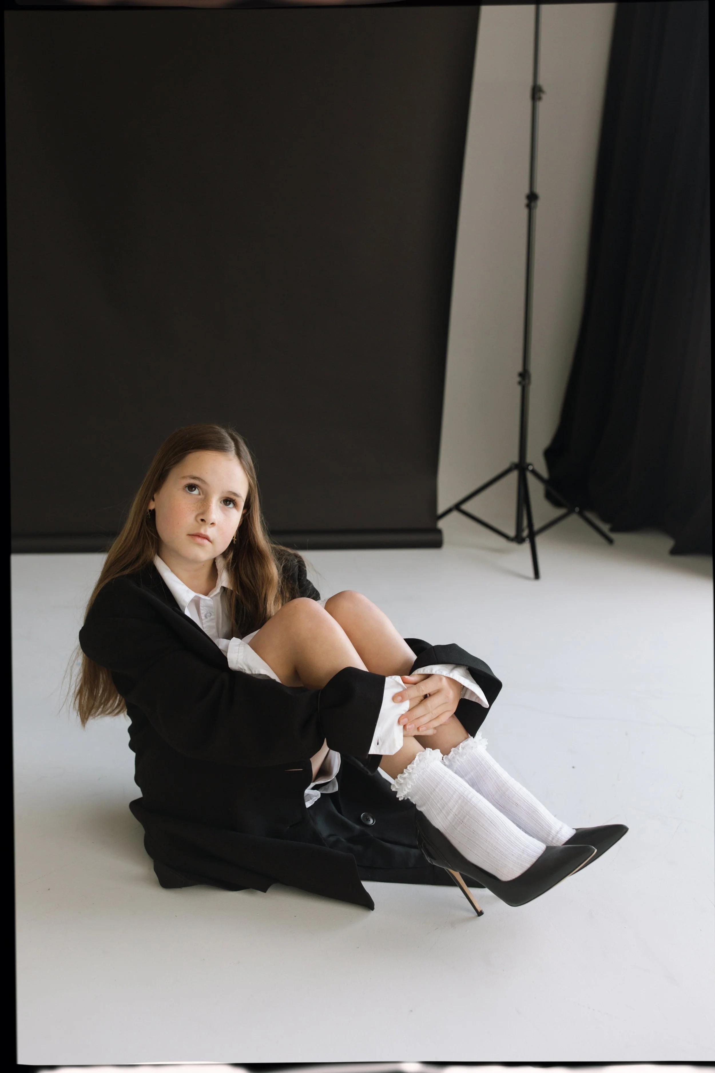 A young girl with long brown hair sitting on the floor in a photography studio, wearing a black blazer, white shirt, white socks, and black high heels, with a black backdrop and studio lighting equipment in the background.