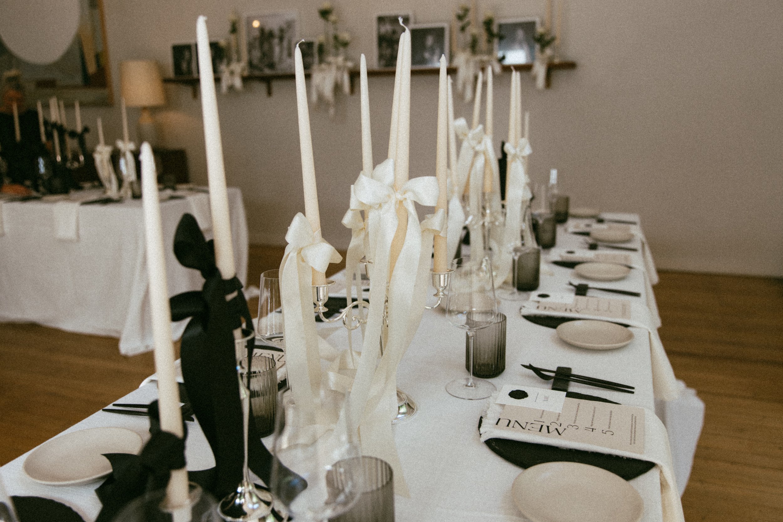 A formal banquet table set with white tablecloths, tall white candles in candelabras with cream ribbons, white plates, and black napkins with chopsticks, in a decorated room with framed photos and decor on the wall.
