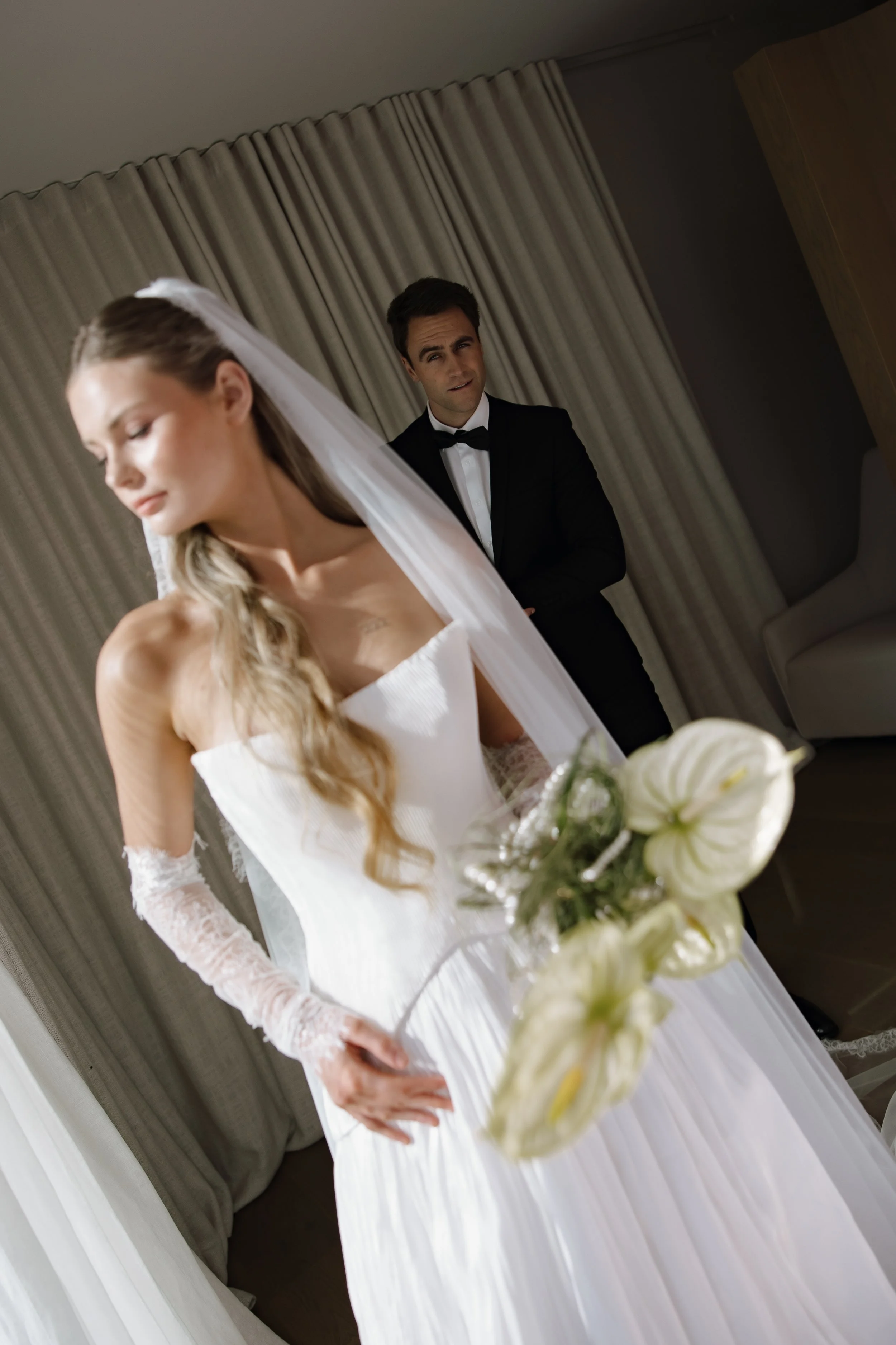 A bride in a white wedding dress holding a bouquet of white flowers, with a groom in a black tuxedo and bow tie standing behind her, in a room with beige curtains.