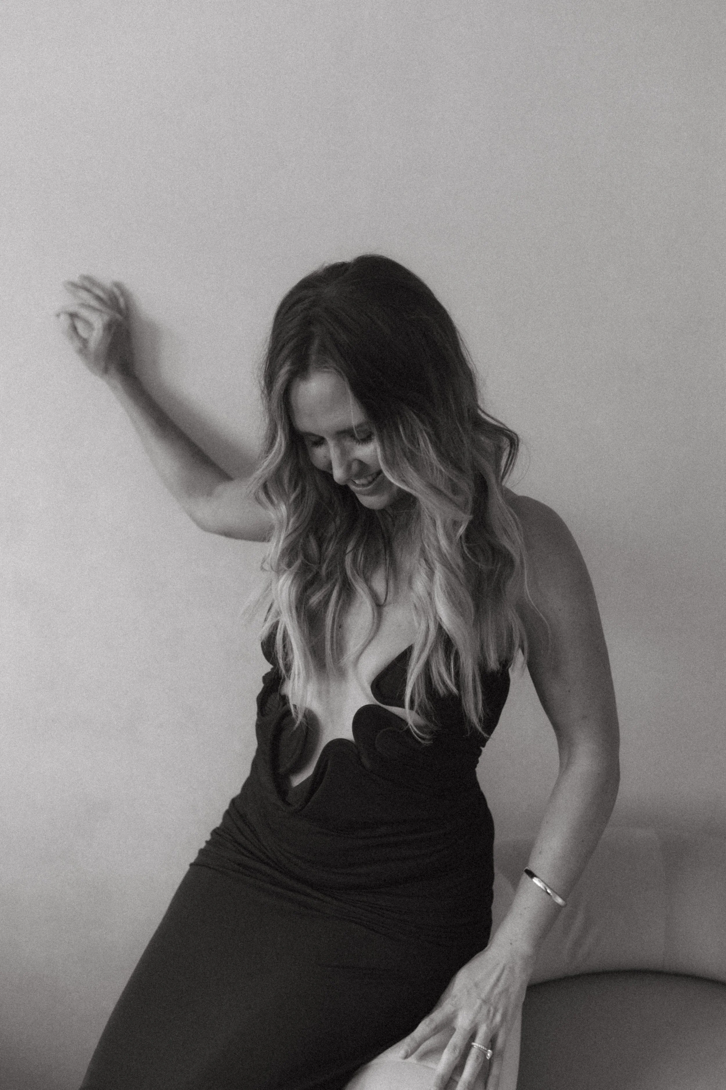 A woman with wavy hair wearing a black dress, smiling and looking down, standing against a plain wall.