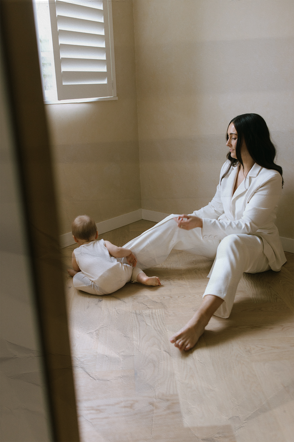 A woman in a white suit sitting on the floor, playing with a baby who is sitting on the floor near her. They are in a room with a window and beige walls.