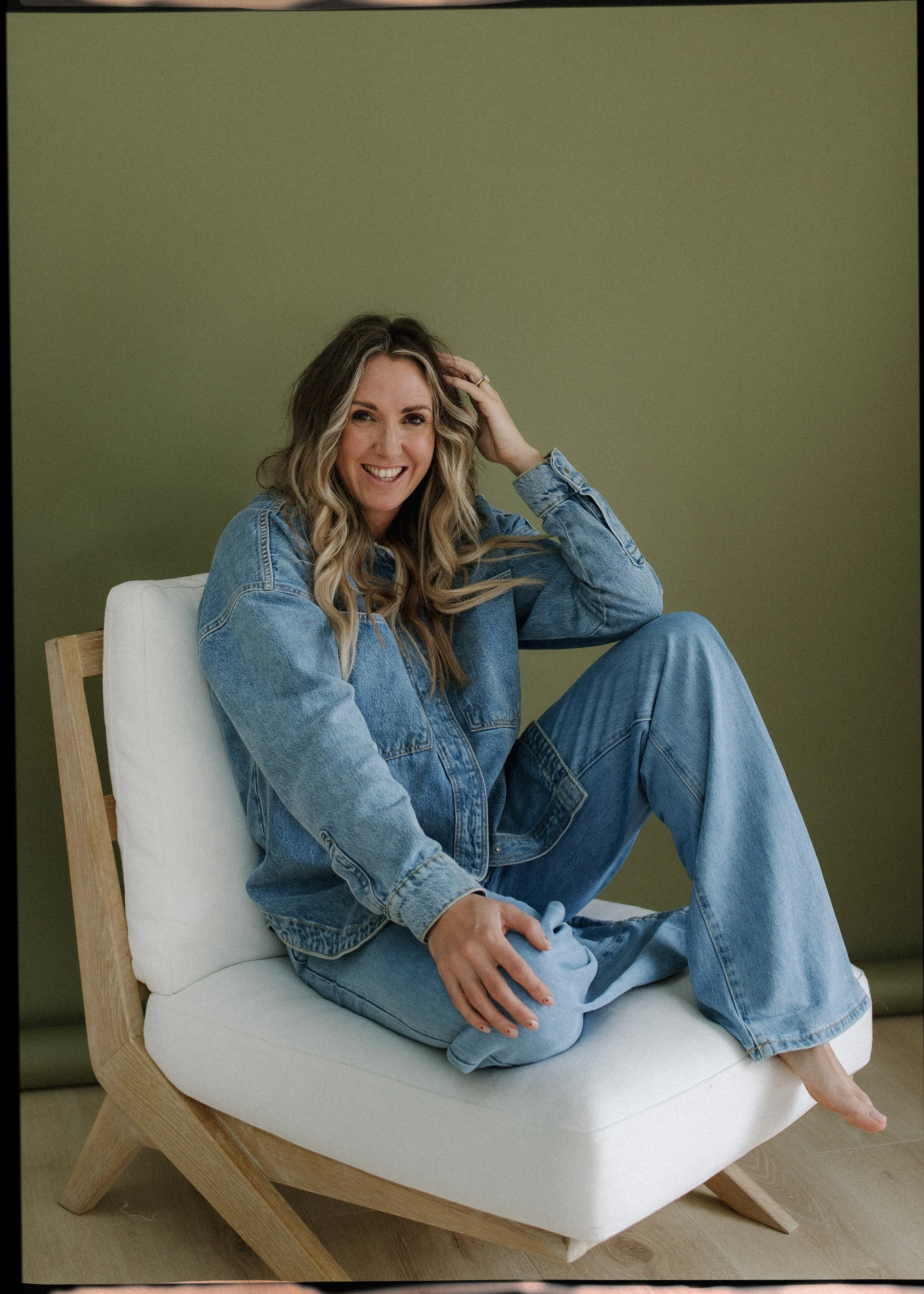 Woman sitting on a white chair with a green background, wearing a denim jacket and jeans, smiling and looking at the camera.