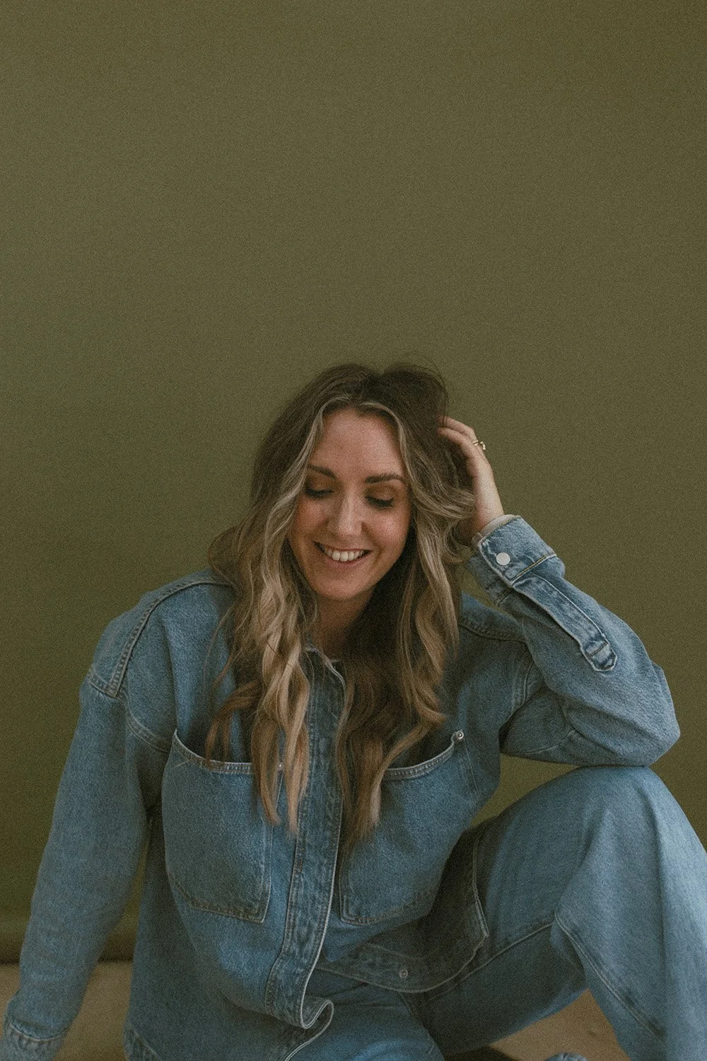 A woman with long wavy hair wearing a denim jacket and jeans sitting on the floor, smiling with her eyes closed, touching her hair, in front of a plain olive green background.