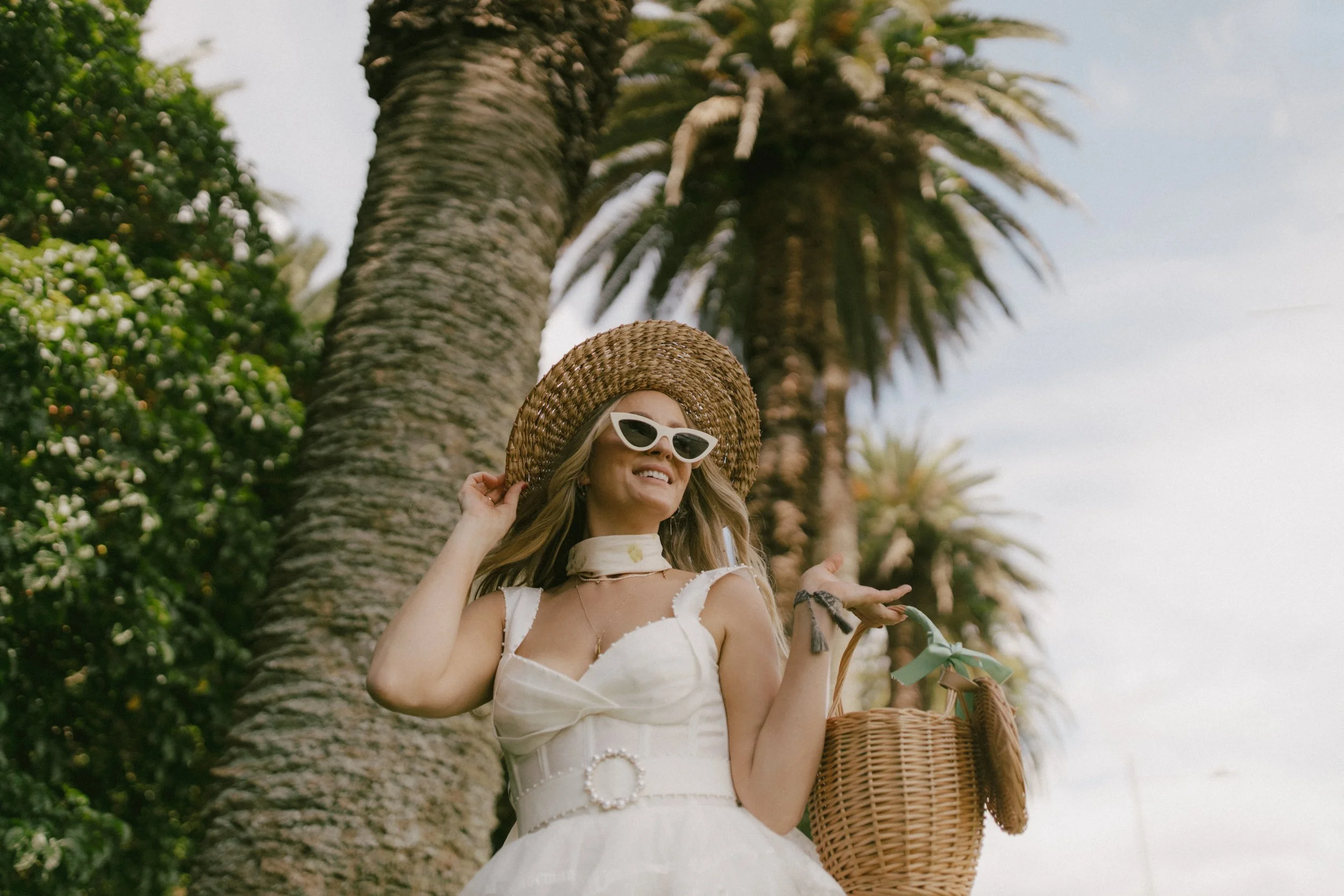 A woman in a white dress with a large straw hat and white sunglasses standing outdoors near palm trees, smiling, holding a wicker picnic basket.