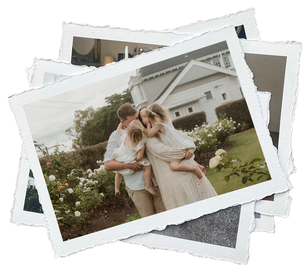 Family of four, two adults and two young girls, embracing and kissing outdoors near a garden with white and pink roses, in front of a white house with a gray roof.