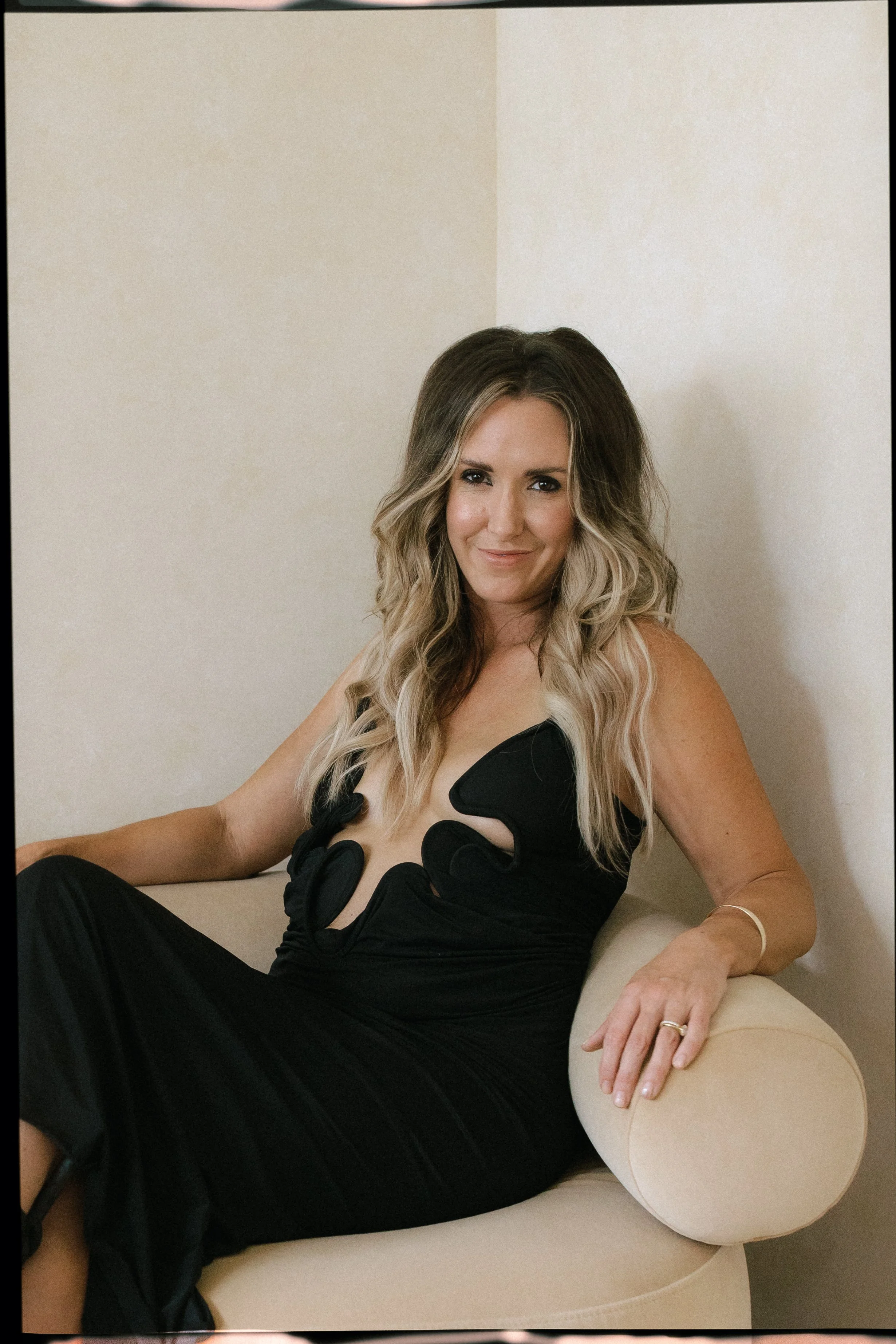 A woman with wavy, blonde hair sitting comfortably on a beige sofa, wearing a black dress with cut-out details, and smiling slightly at the camera.