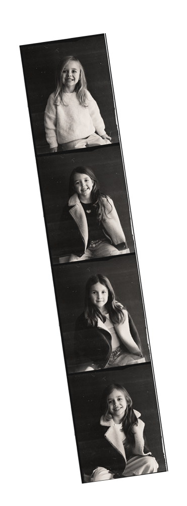 Black and white photo strip showing four pictures of a young girl with long hair, smiling in each photo, wearing a light-colored sweater and jacket, sitting against a dark background.
