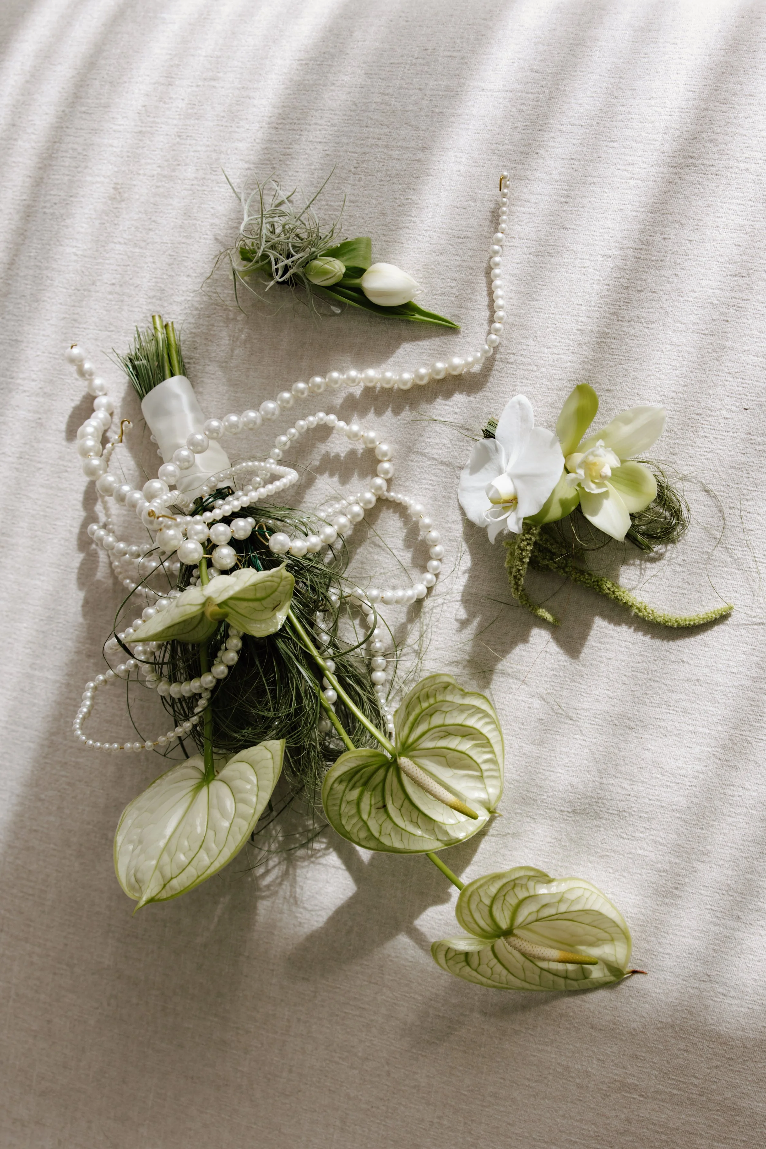 Elegant floral arrangement with white flowers, green leaves, pearls, and greenery arranged on a light fabric background.