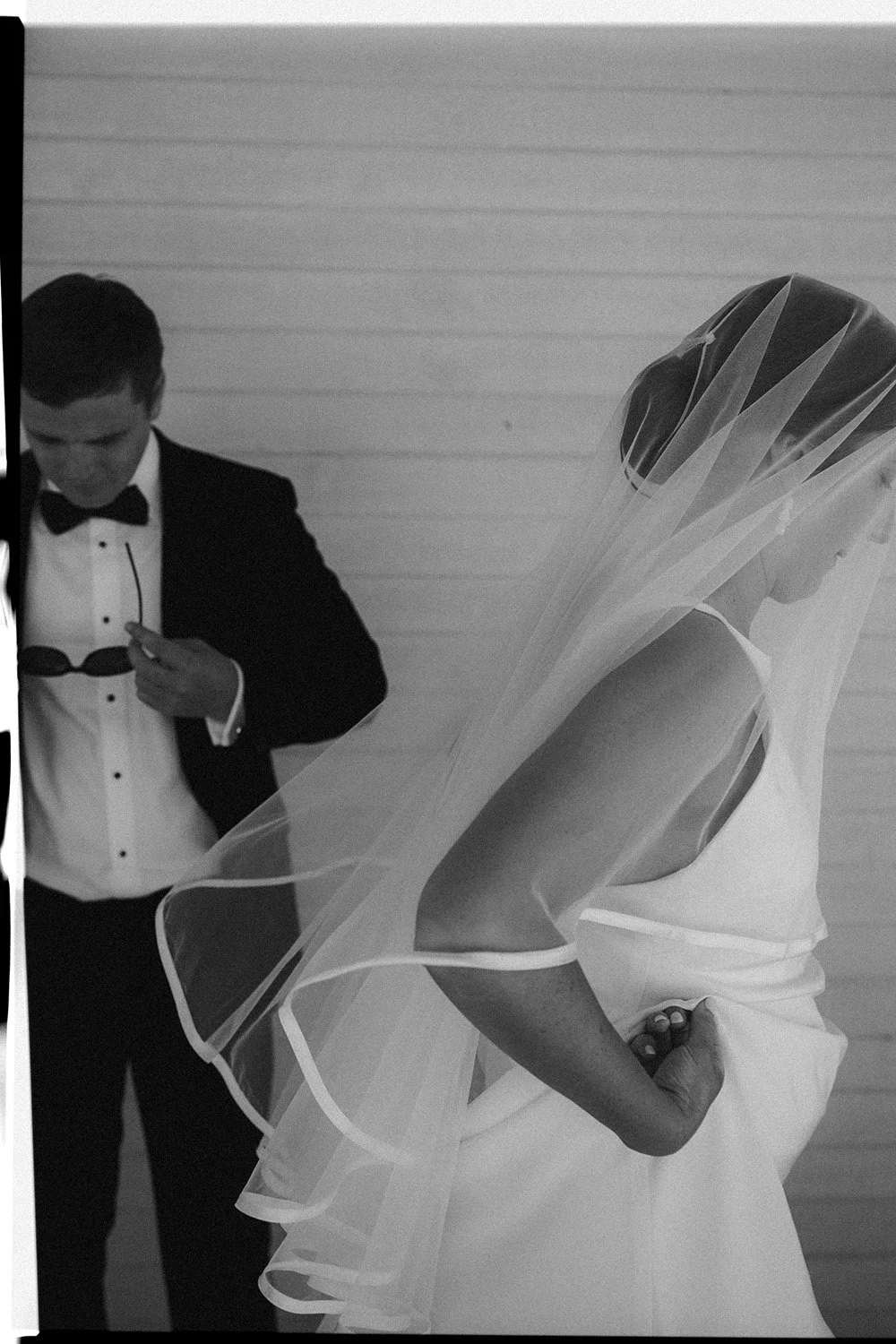 A bride in a sleeveless white wedding dress with a veil covering her face and a man in a tuxedo with a bowtie, standing against a plain wall. The bride's hand is resting on her hip.
