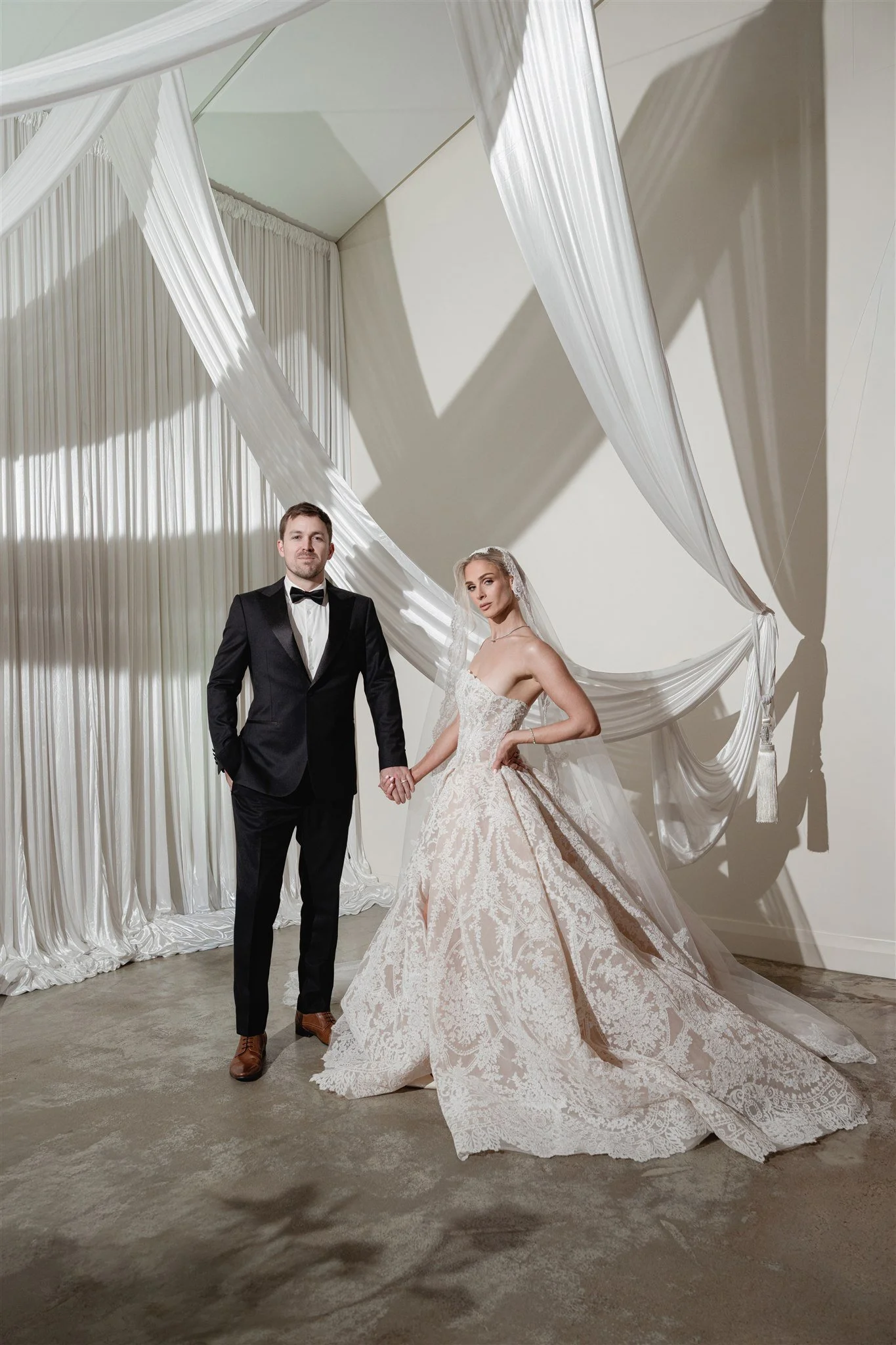 A bride and groom dressed in wedding attire standing hand in hand in front of white drapery and shadows.