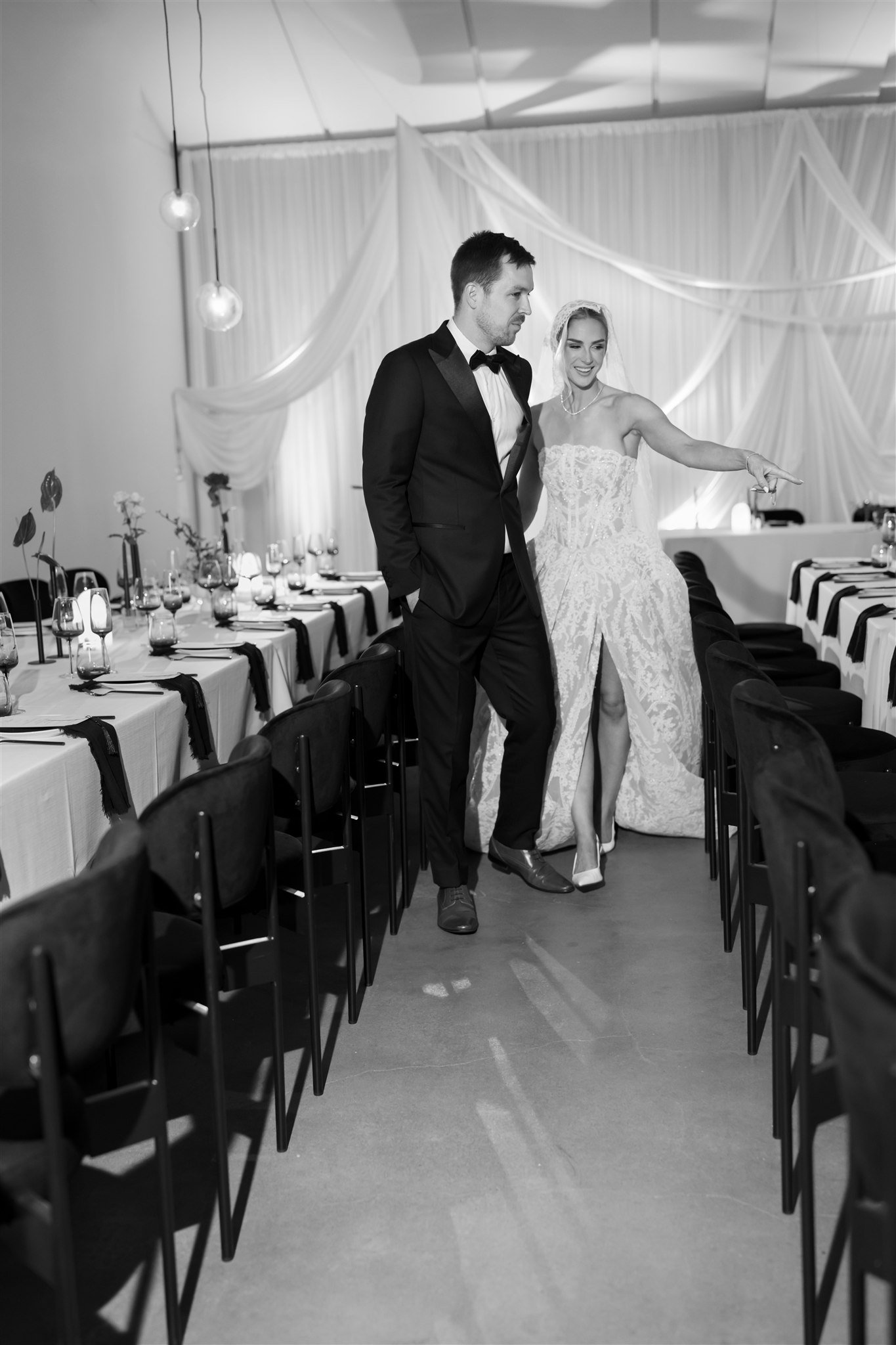 A bride and groom dressed in wedding attire walking through a decorated banquet hall, with the bride pointing towards something off-camera.